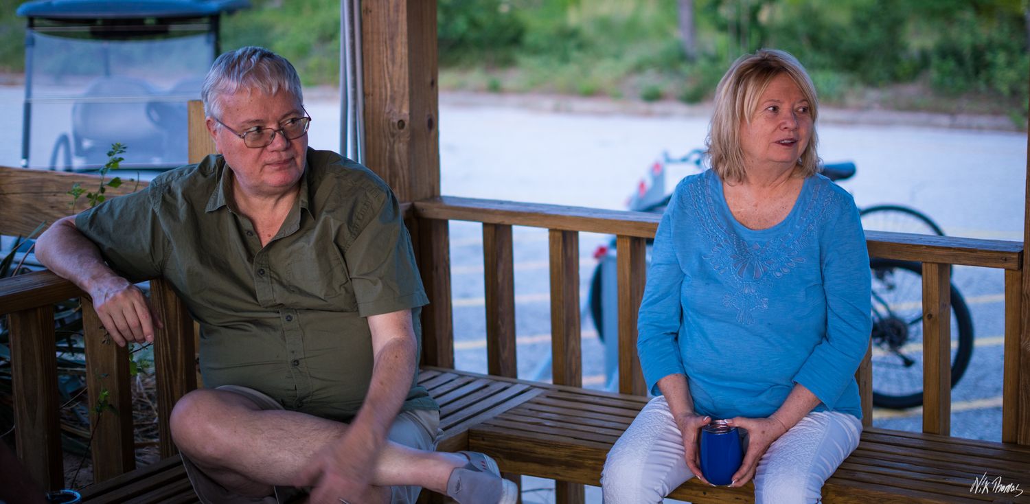 An elderly couple sitting on a wooden bench on a porch, man in green shirt and woman in blue sweater.