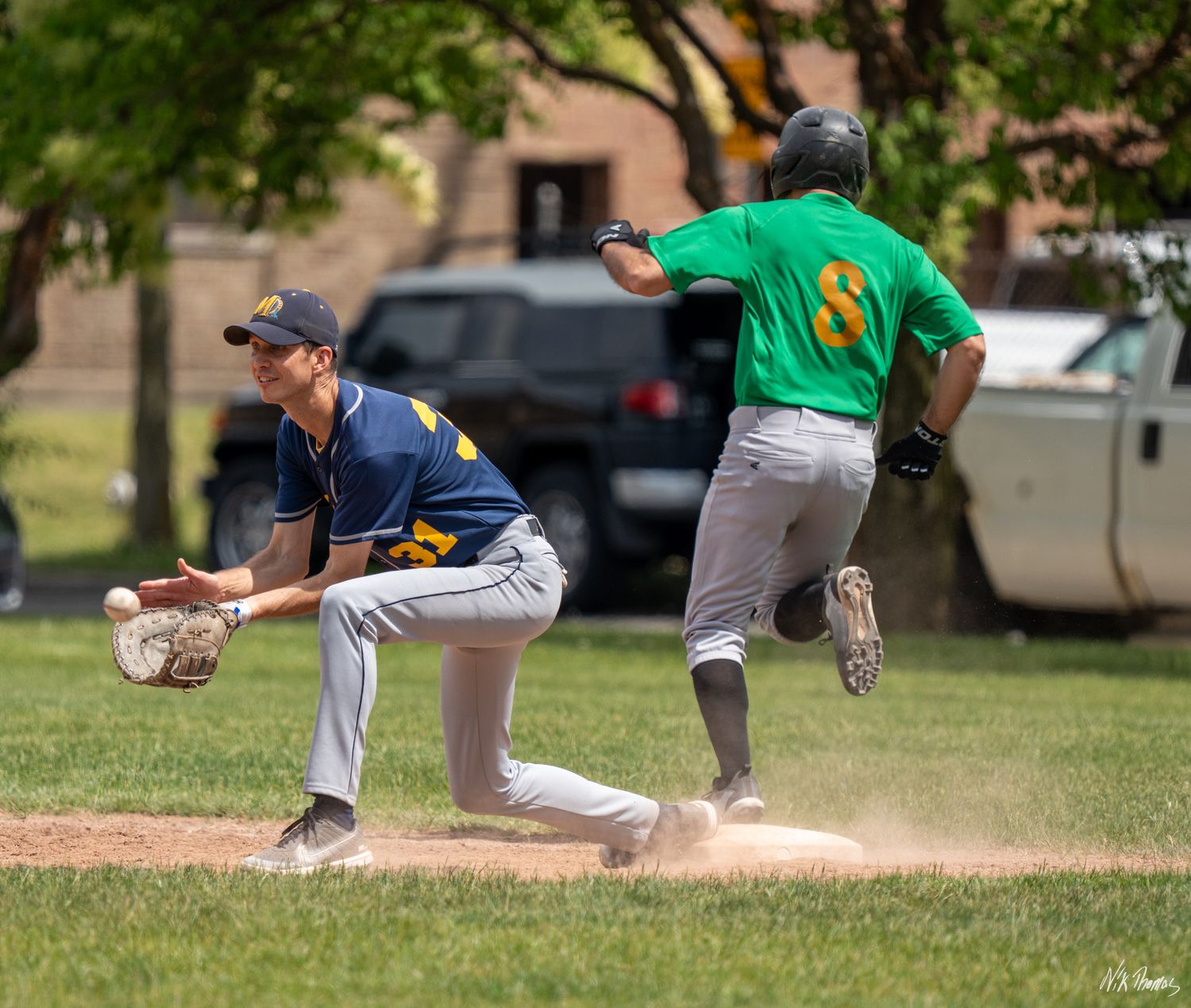 Baseball player in green jersey runs to base as fielder in navy uniform crouches to catch ball during sunny outdoor game.