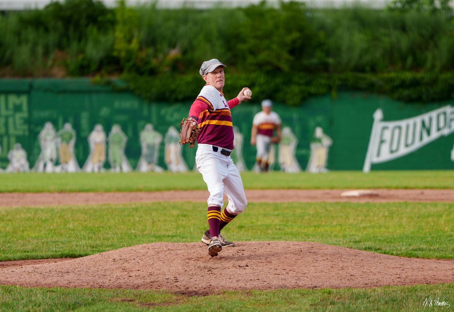 Youth baseball pitcher in maroon and gold uniform releases a pitch from the mound during a game.