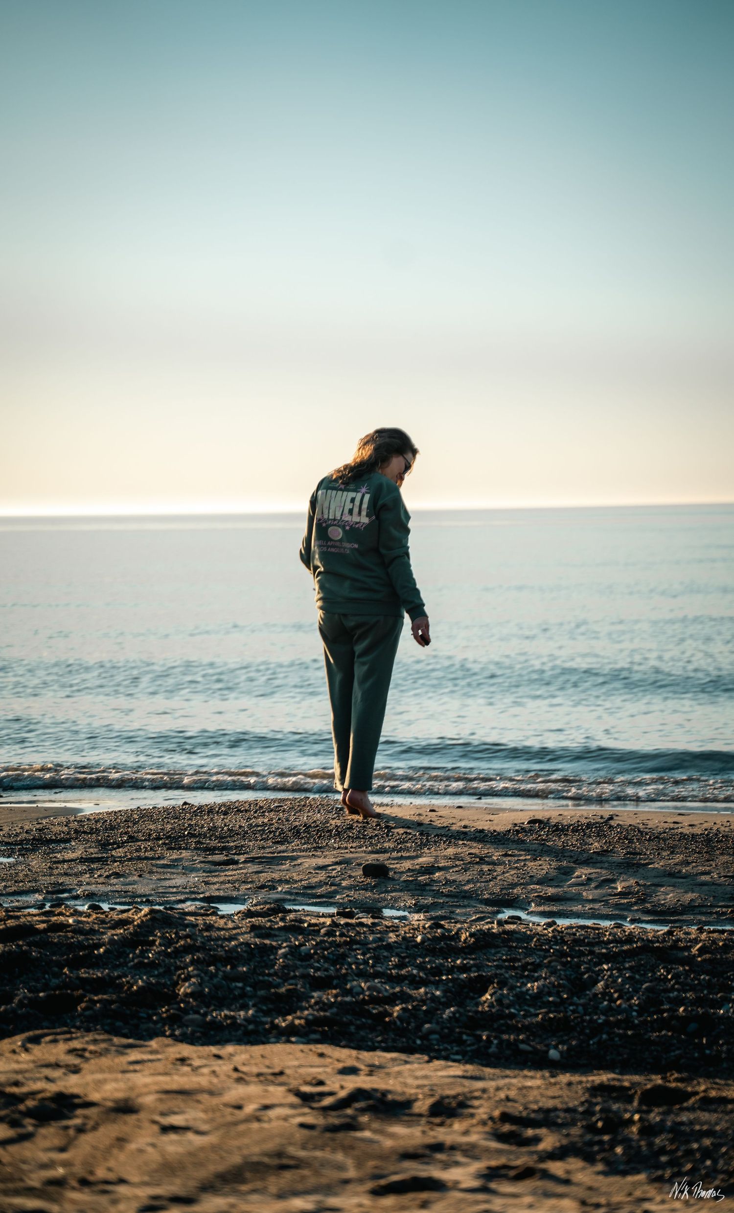 A woman in a dark Powell hoodie stands barefoot on a rocky beach, gazing down at the shore with calm ocean behind her.