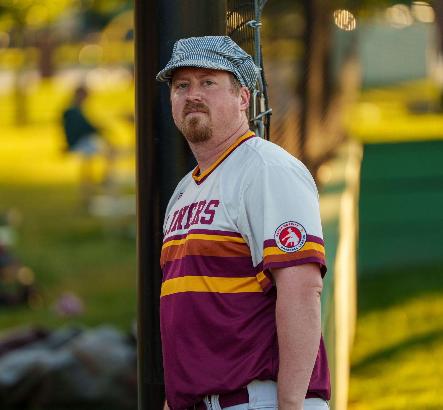 Man in vintage baseball uniform leaning against pole on sunny baseball field.