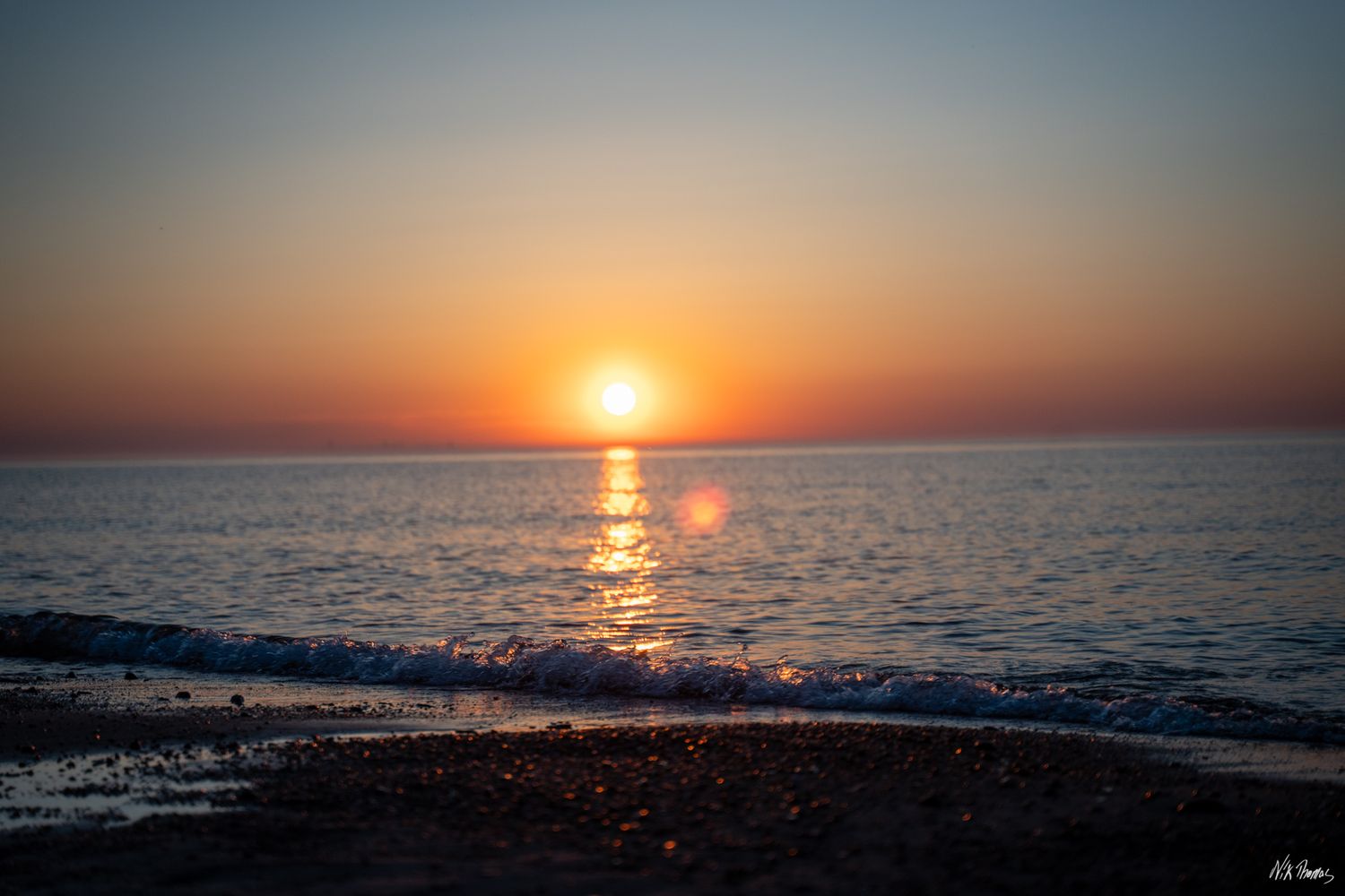 Stunning ocean sunset with vibrant orange sky reflecting on calm waves gently washing over a rocky beach.