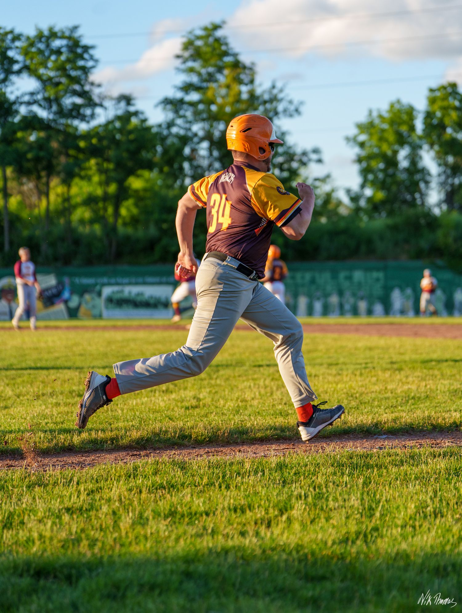 A youth baseball player in a yellow jersey and orange helmet sprints across a green field during a sunny evening game.