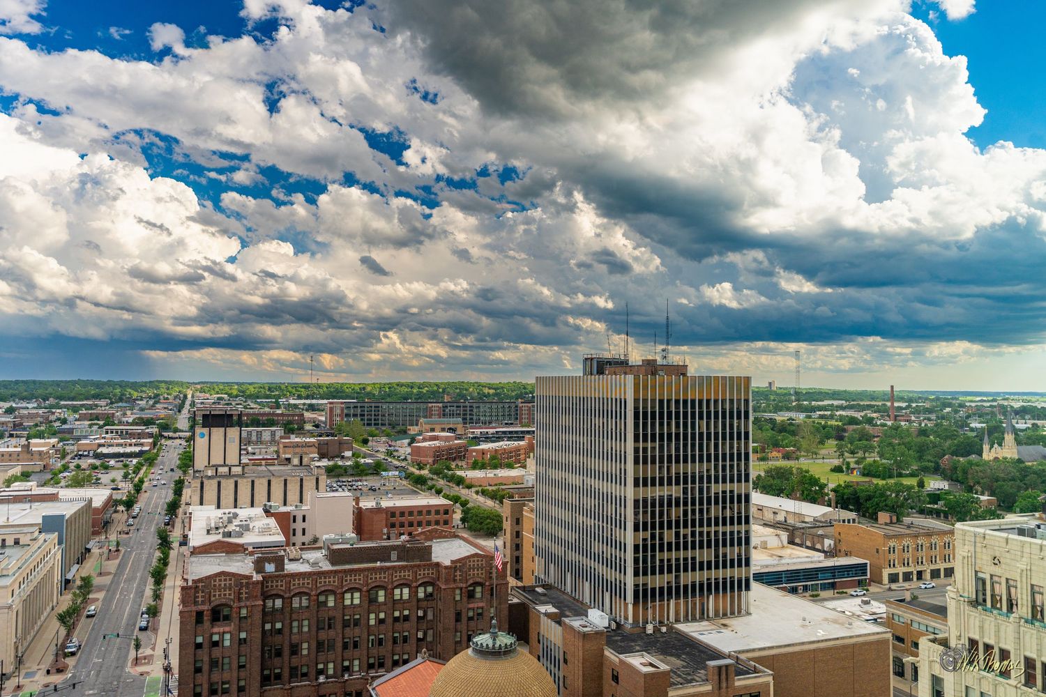 Aerial view of a city skyline under dramatic blue sky with large white clouds, featuring tall buildings and urban streets.