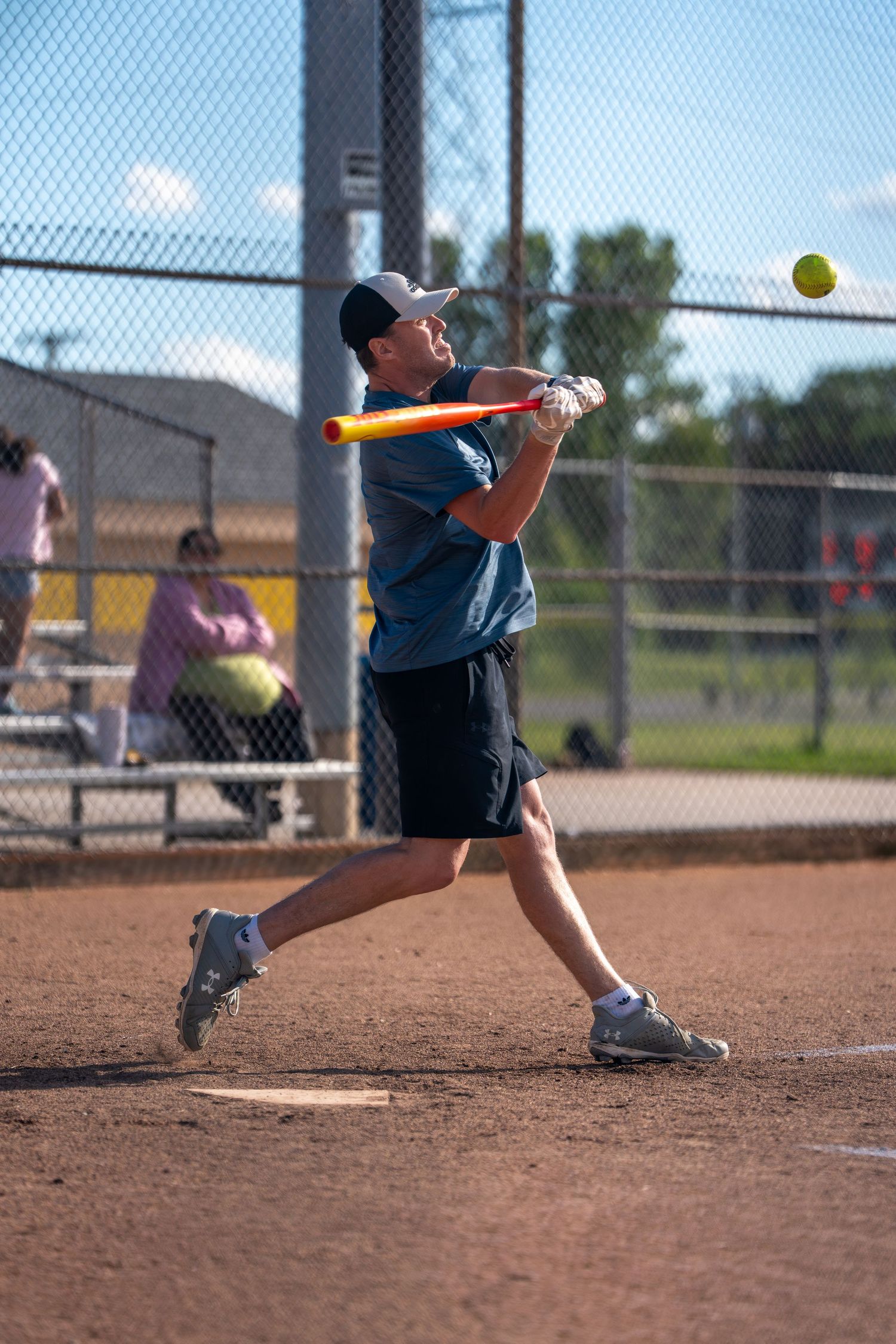 Man in blue shirt swinging an orange bat at a yellow softball on a dirt field during a recreational game.