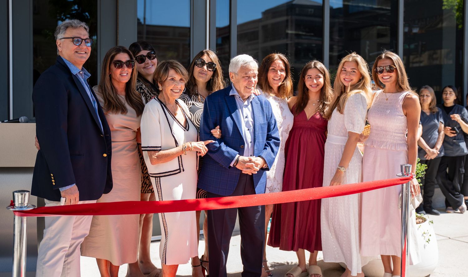Group of well-dressed people posing together at an outdoor ribbon-cutting ceremony on a sunny day.