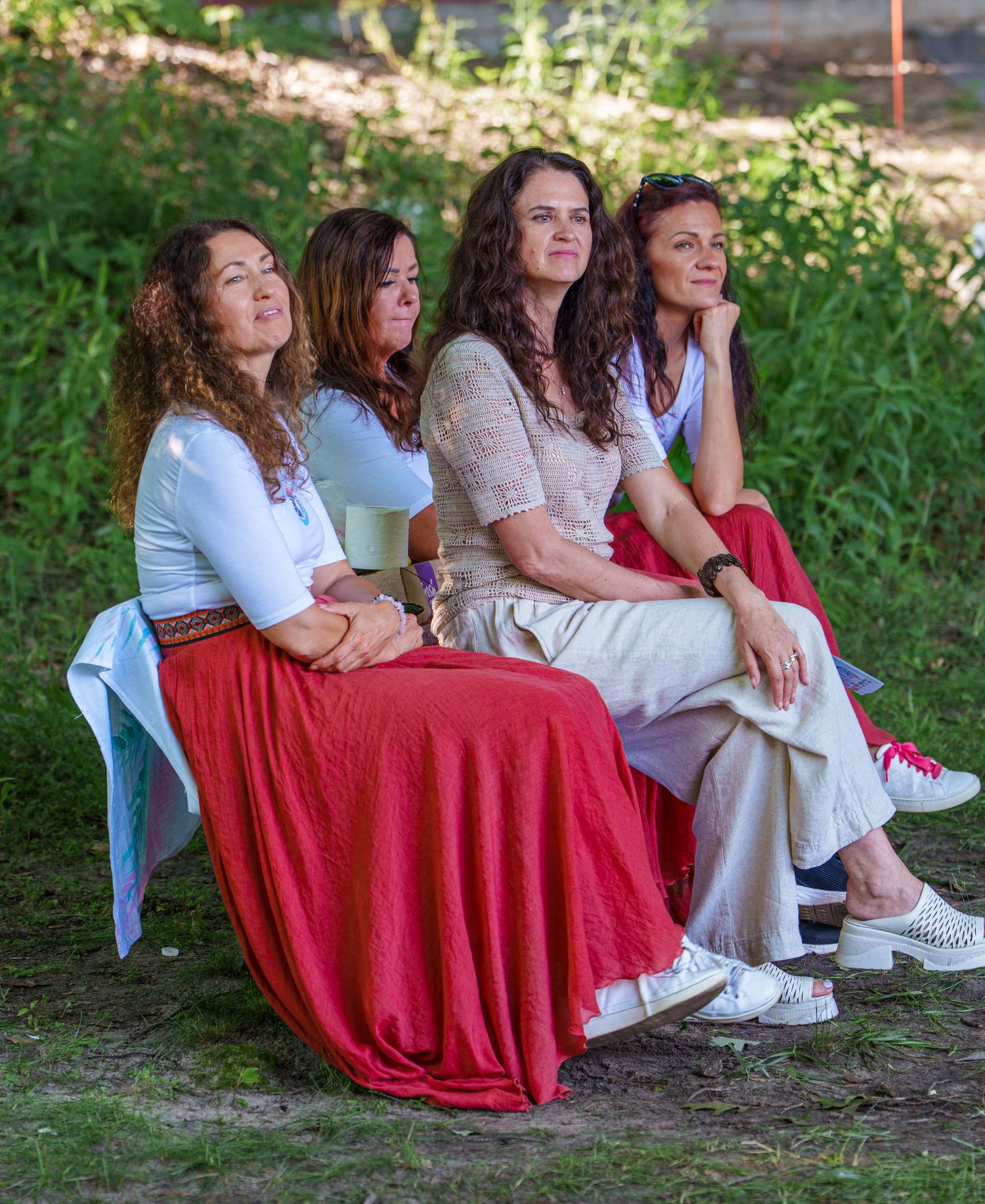 Four women sitting together outdoors in flowing red and white skirts, enjoying a sunny day in a garden setting.