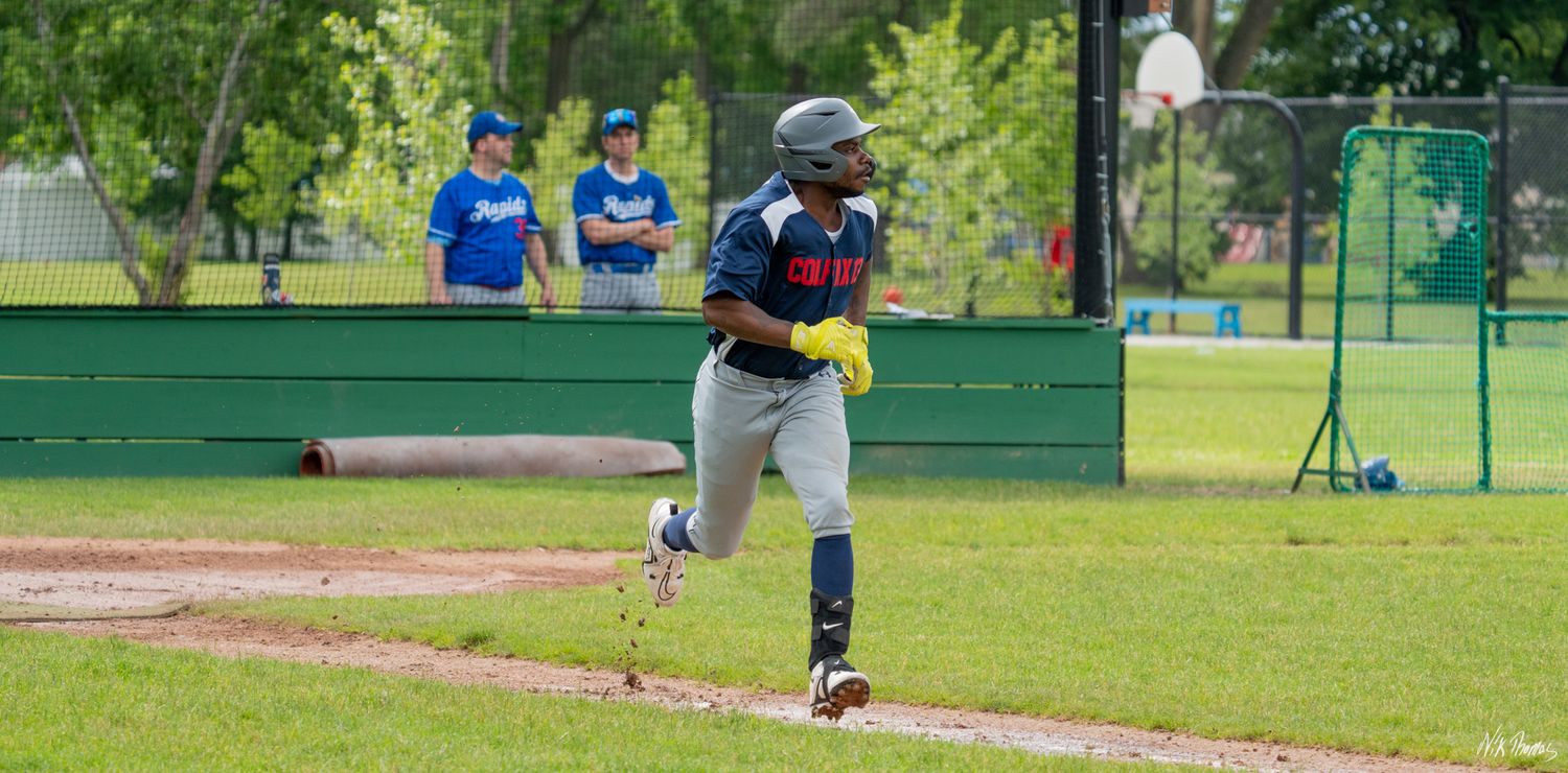 Youth baseball player runs the bases in a game, wearing a helmet and yellow batting gloves, with coaches watching.