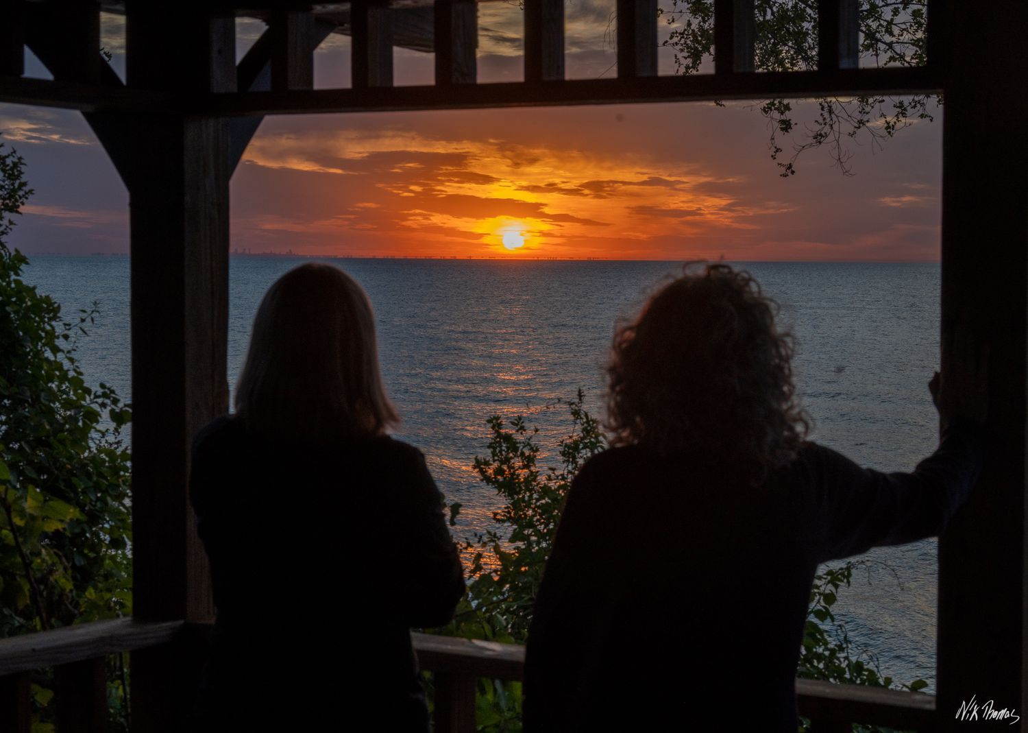 Two silhouetted figures watch a vibrant orange sunset over a calm ocean from a wooden porch.