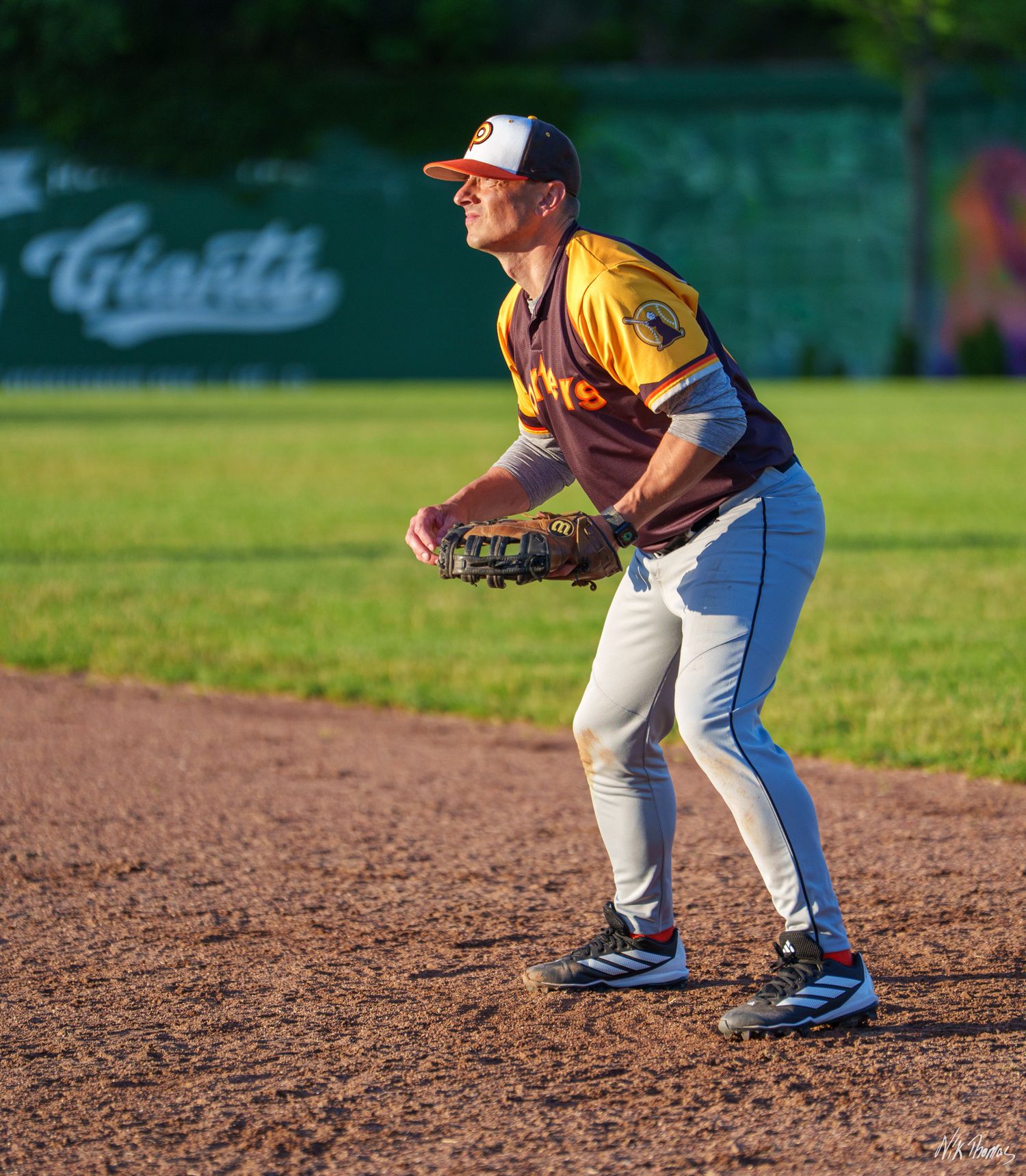 Baseball infielder in yellow uniform crouches in ready position on dirt infield during evening game.