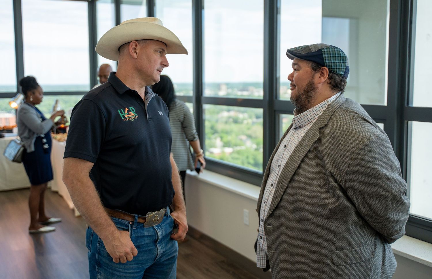 Two men in casual attire having conversation near large windows with city view in background.