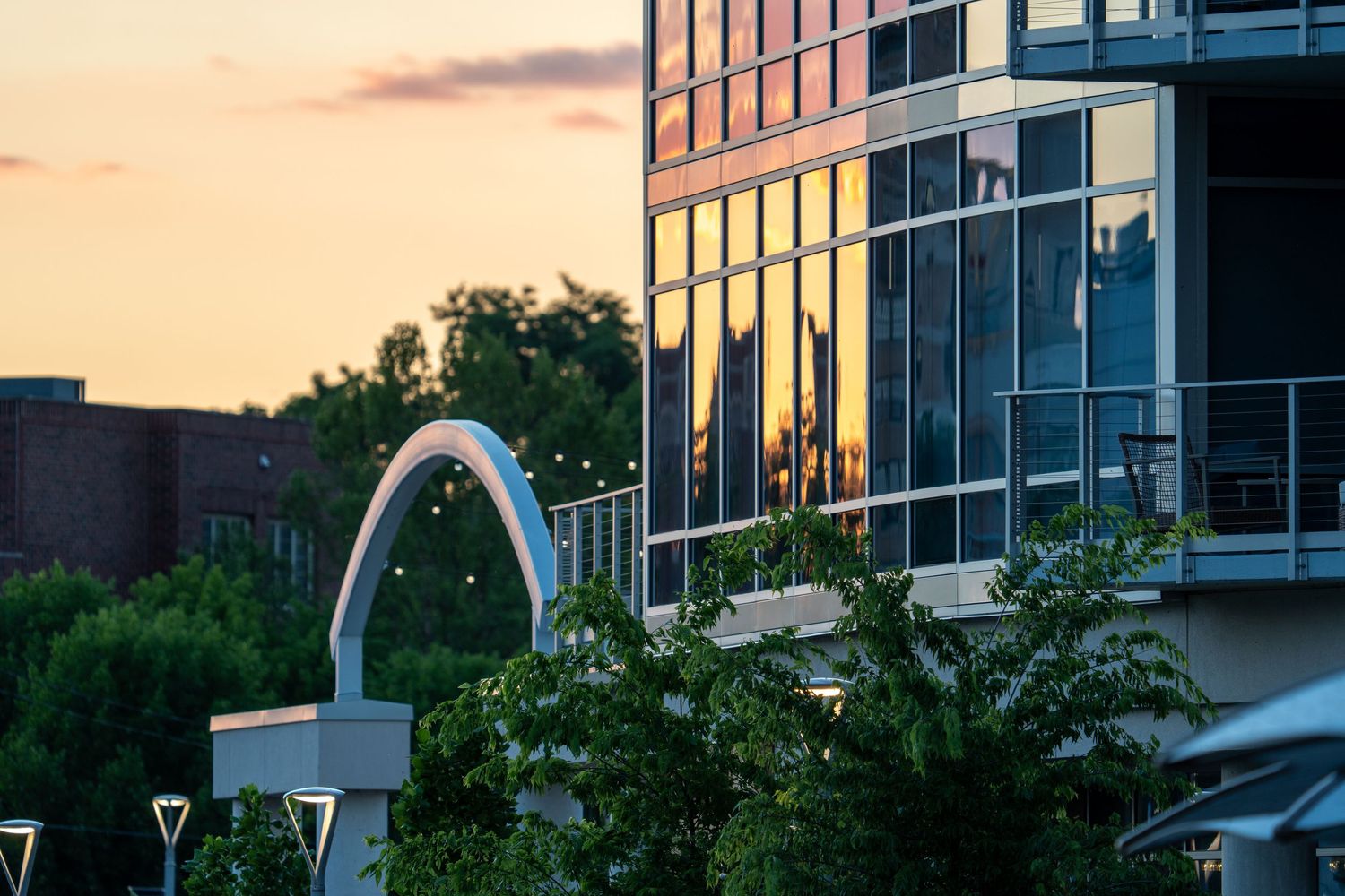 Modern glass building reflects golden sunset light beside an illuminated arch sculpture and green trees.