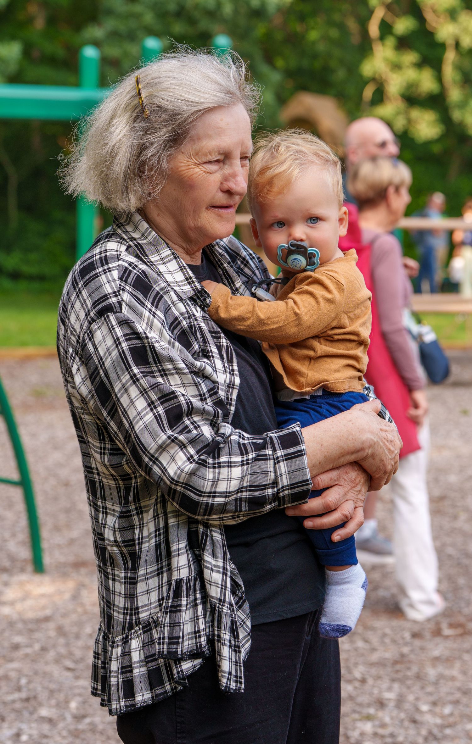 Elderly grandmother with gray hair holding a young toddler with a pacifier at an outdoor playground.