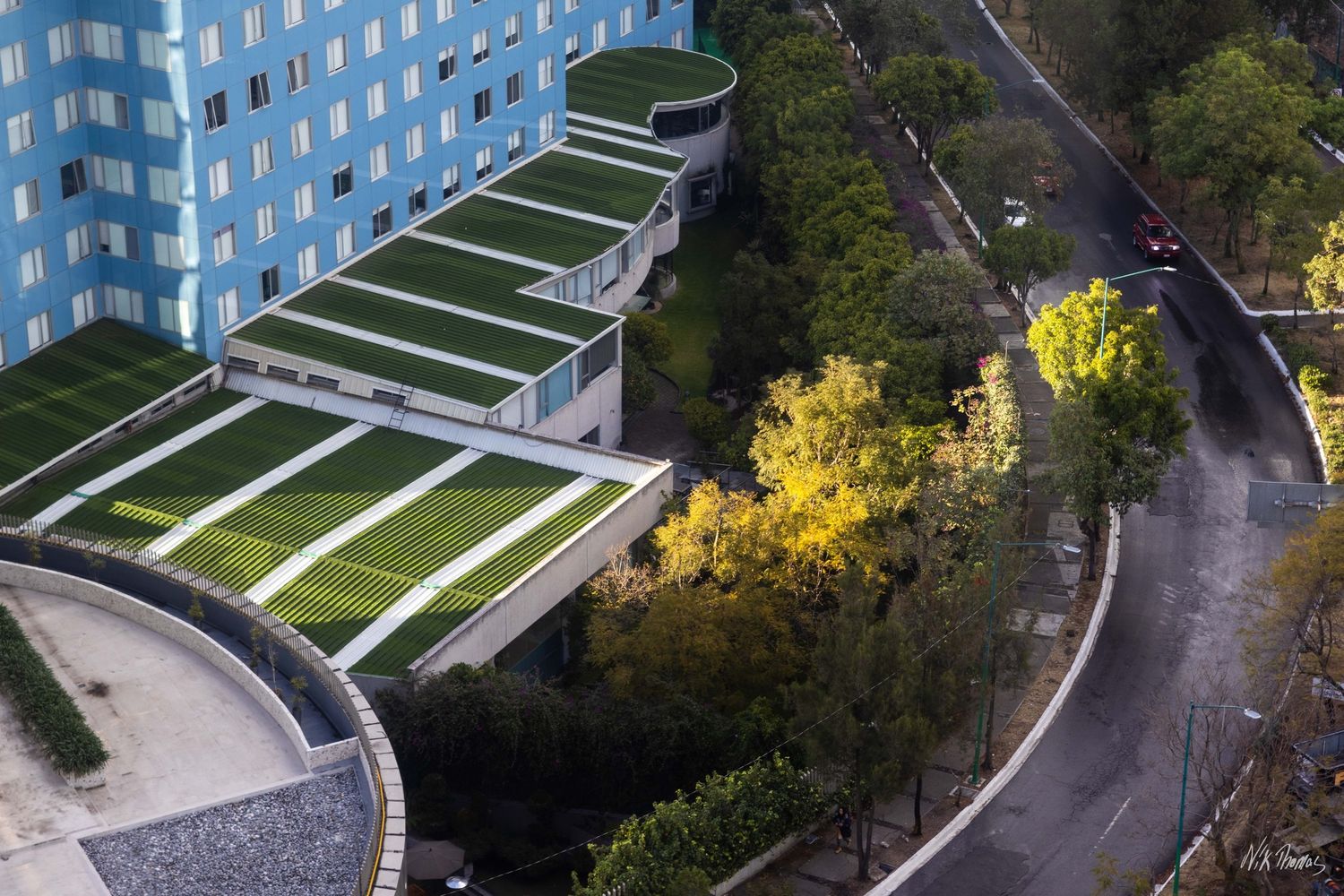 Aerial view of a modern blue building with green rooftop gardens surrounded by tree-lined roads in an urban setting.