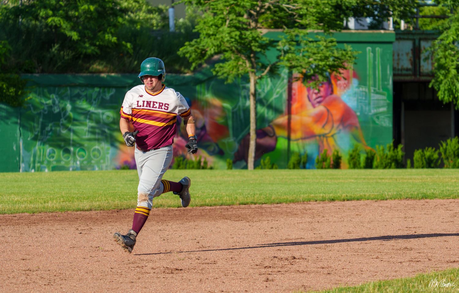 Baseball player in Liners uniform sprinting bases on a dirt field with colorful mural in background.