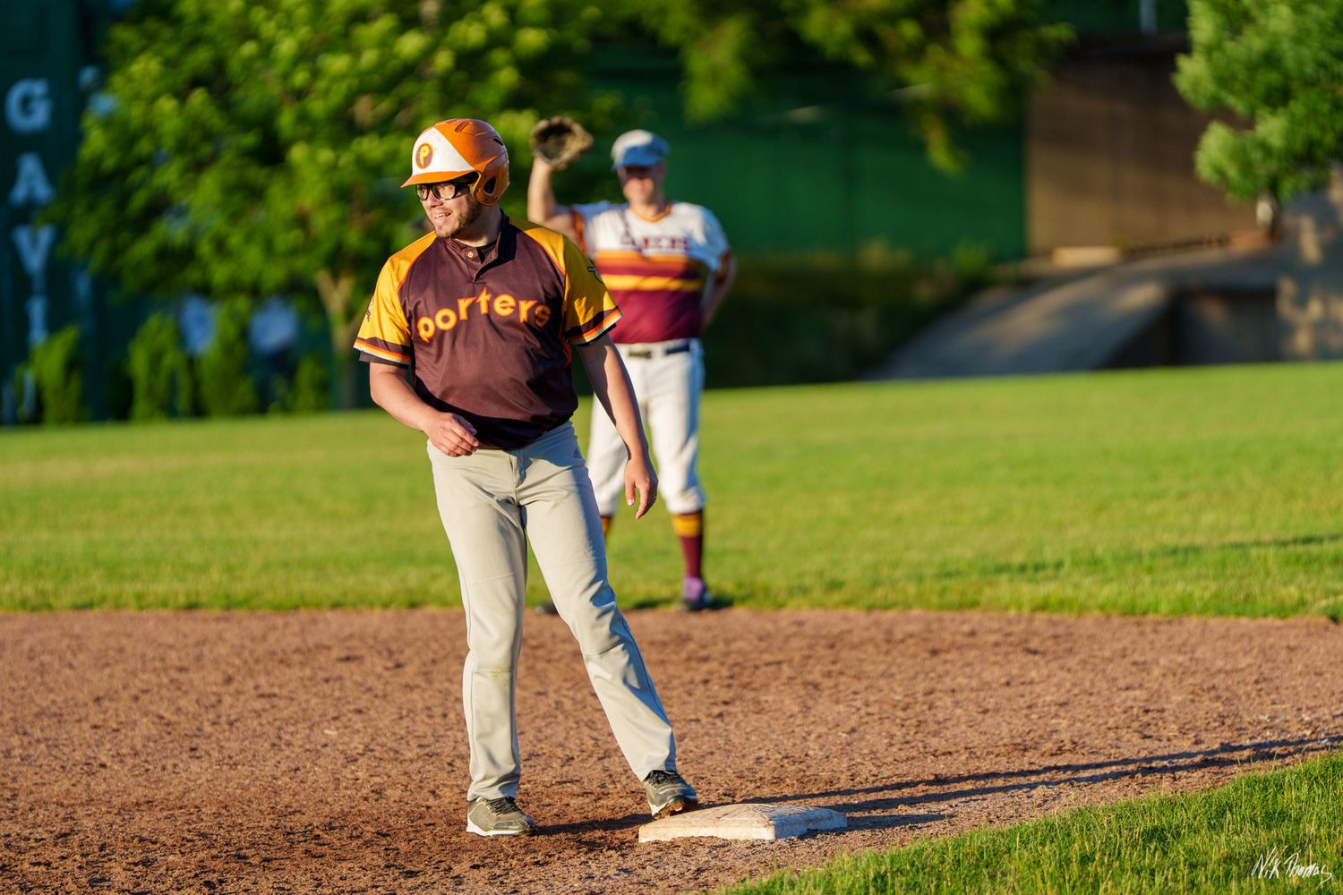 A baseball coach in a Dorfer jersey stands on the baseline as a player pitches in the background.