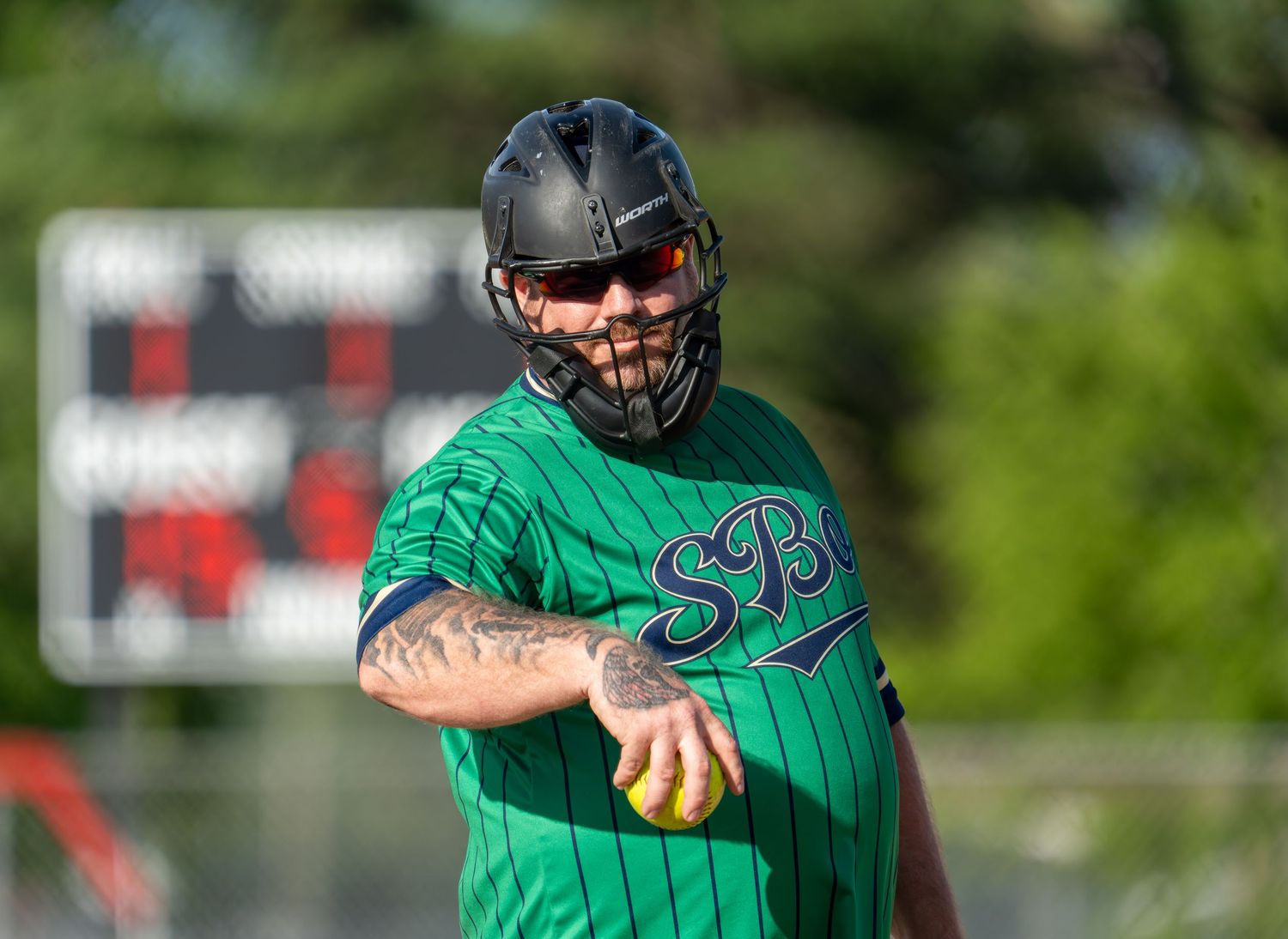 Softball player in green SB jersey and helmet prepares to throw a yellow ball on a sunny day.