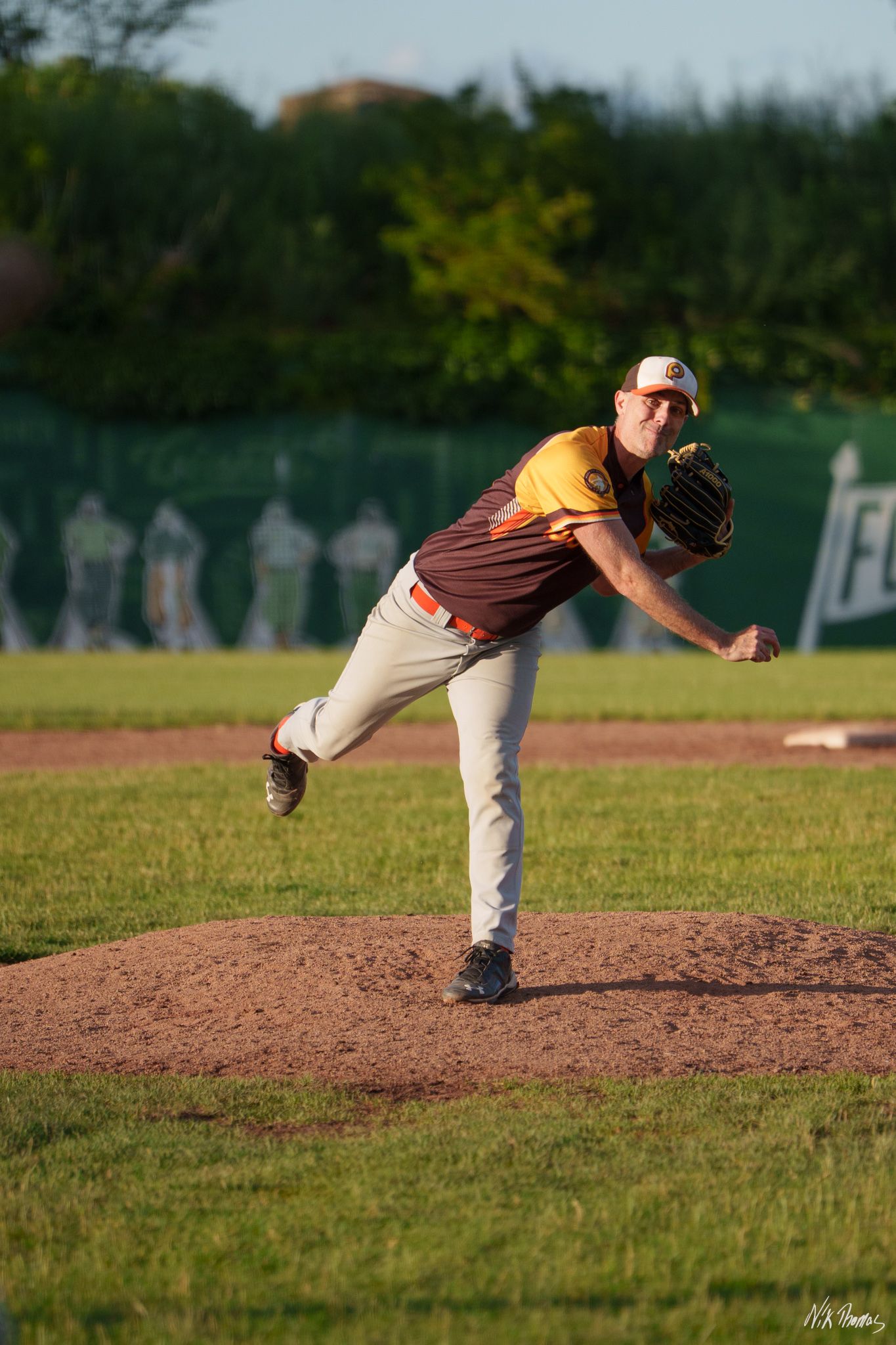 Baseball pitcher in brown and yellow uniform mid-throw on mound during sunny evening game.