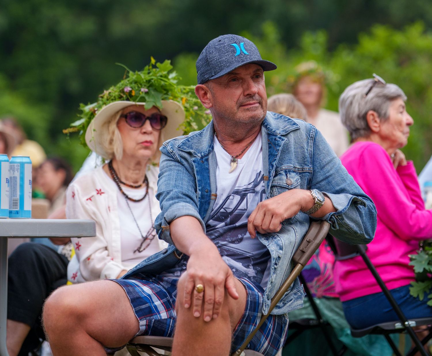 Man in baseball cap and woman wearing floral crown sit outdoors at a summer event among other attendees.