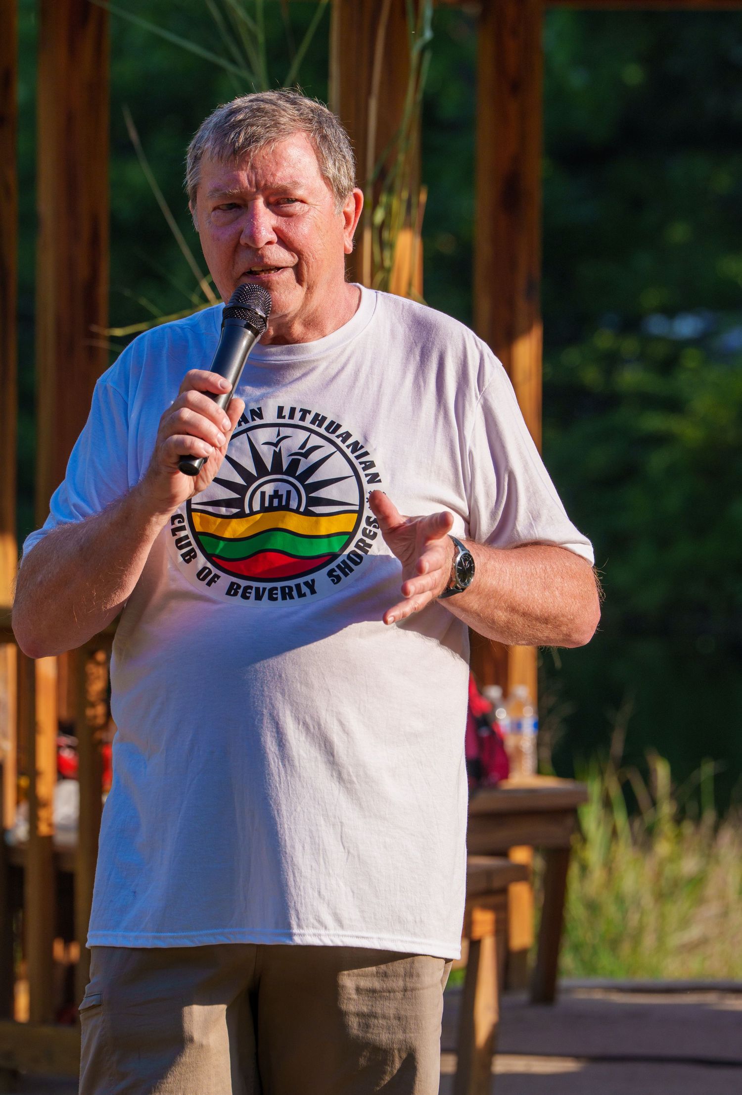 Man speaking into microphone wearing American Lithuanian Club of Beverly Shores t-shirt outdoors.