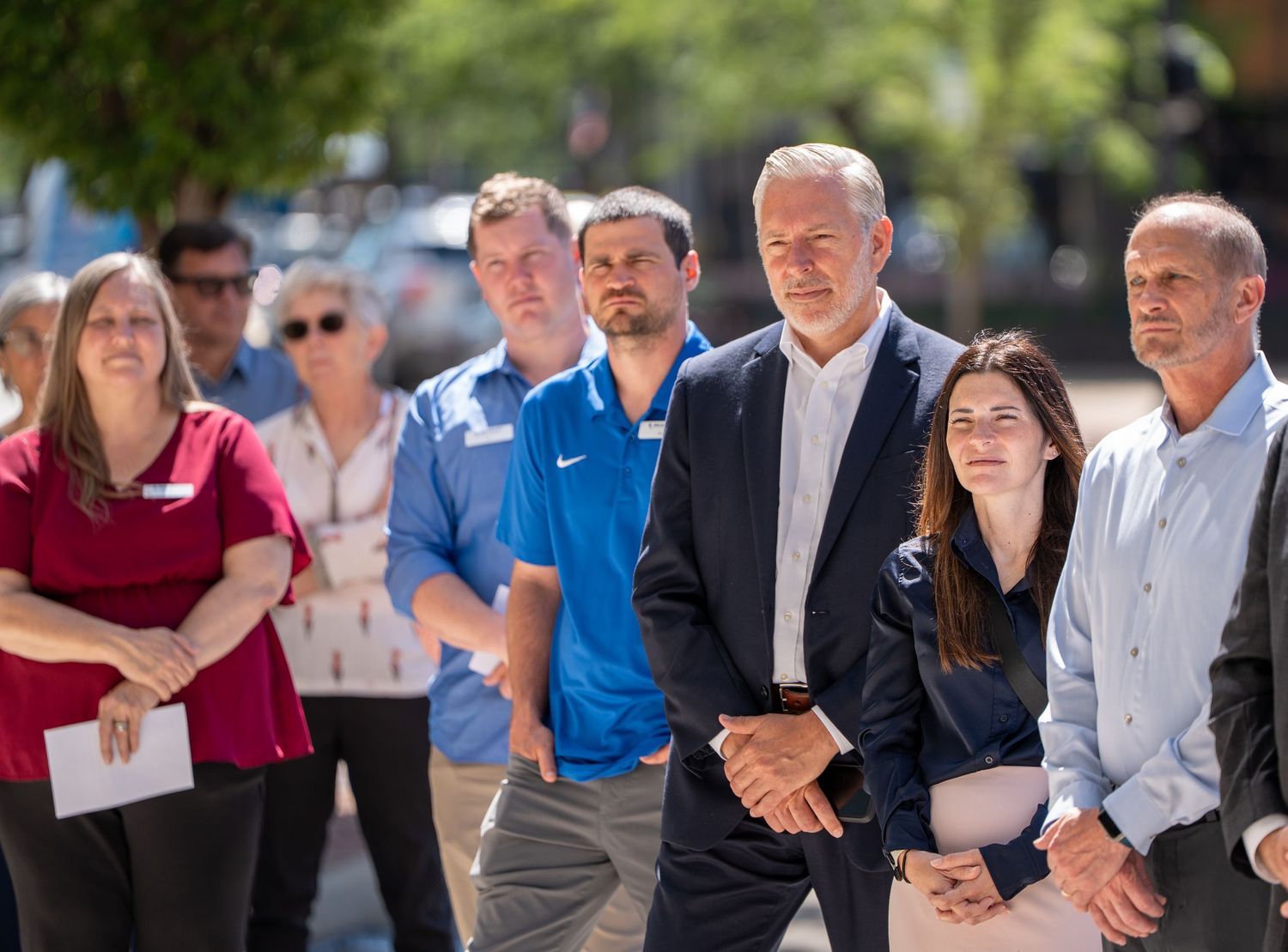 Group of adults standing attentively outdoors at a public community gathering on a sunny day.