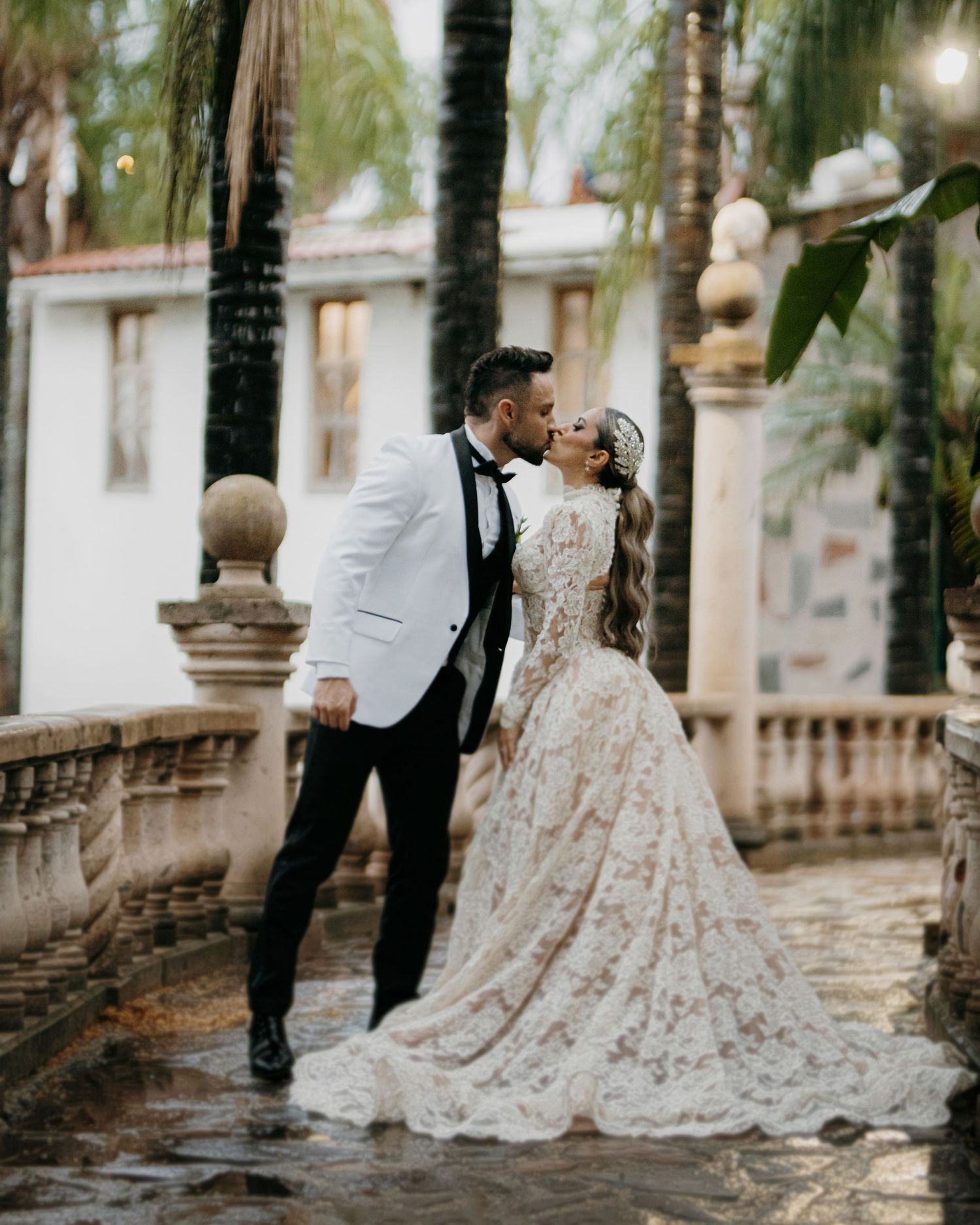 A couple shares a romantic kiss on a stone terrace, bride in lace gown and groom in white tuxedo jacket.