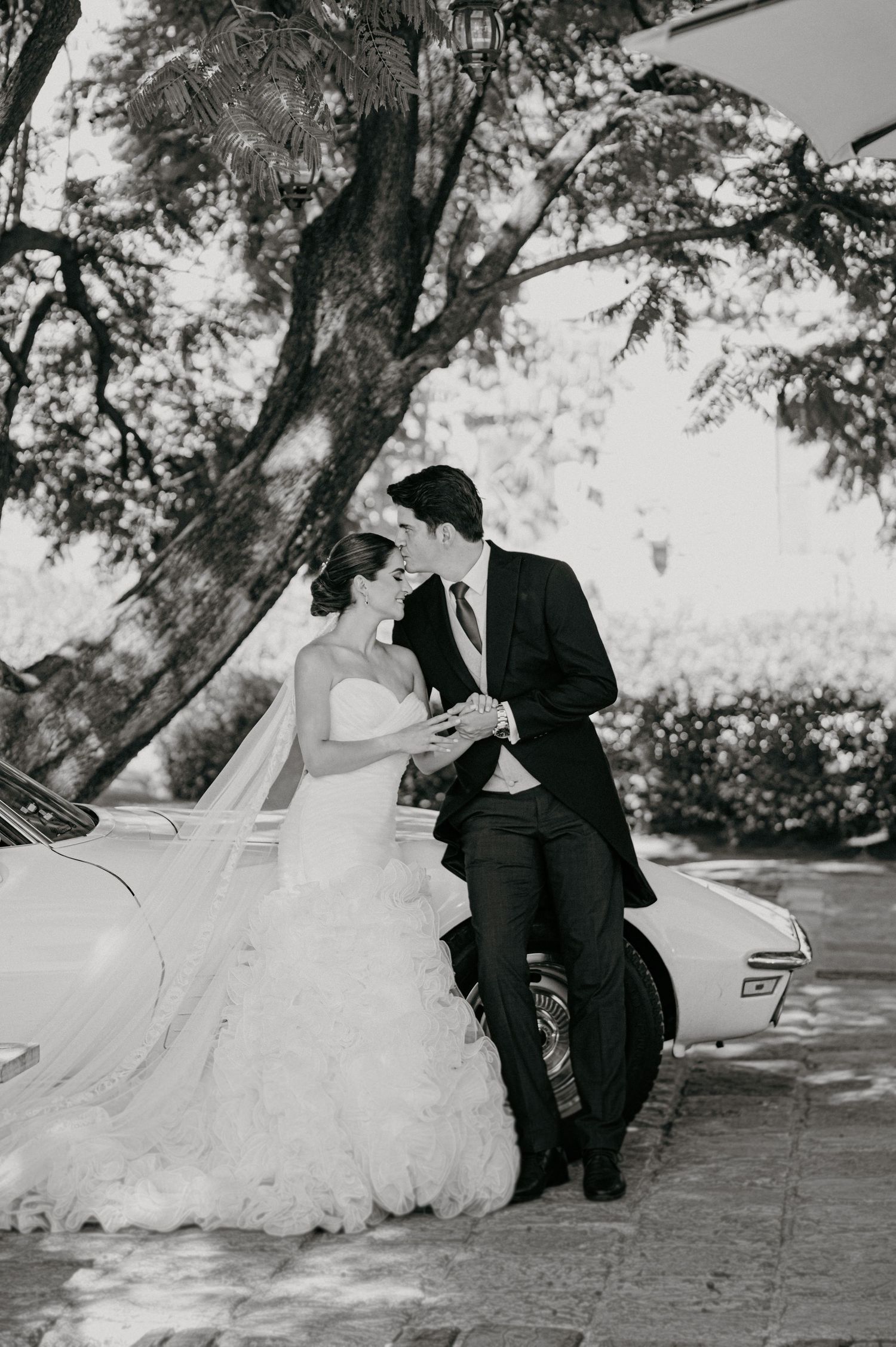 Black and white photo of a bride and groom sharing a romantic moment beside a vintage car outdoors.