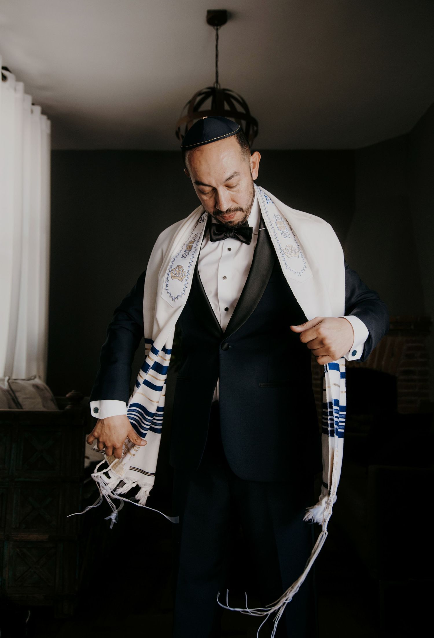 Groom in black tuxedo wearing a Jewish tallit prayer shawl and kippah, preparing for his wedding ceremony.
