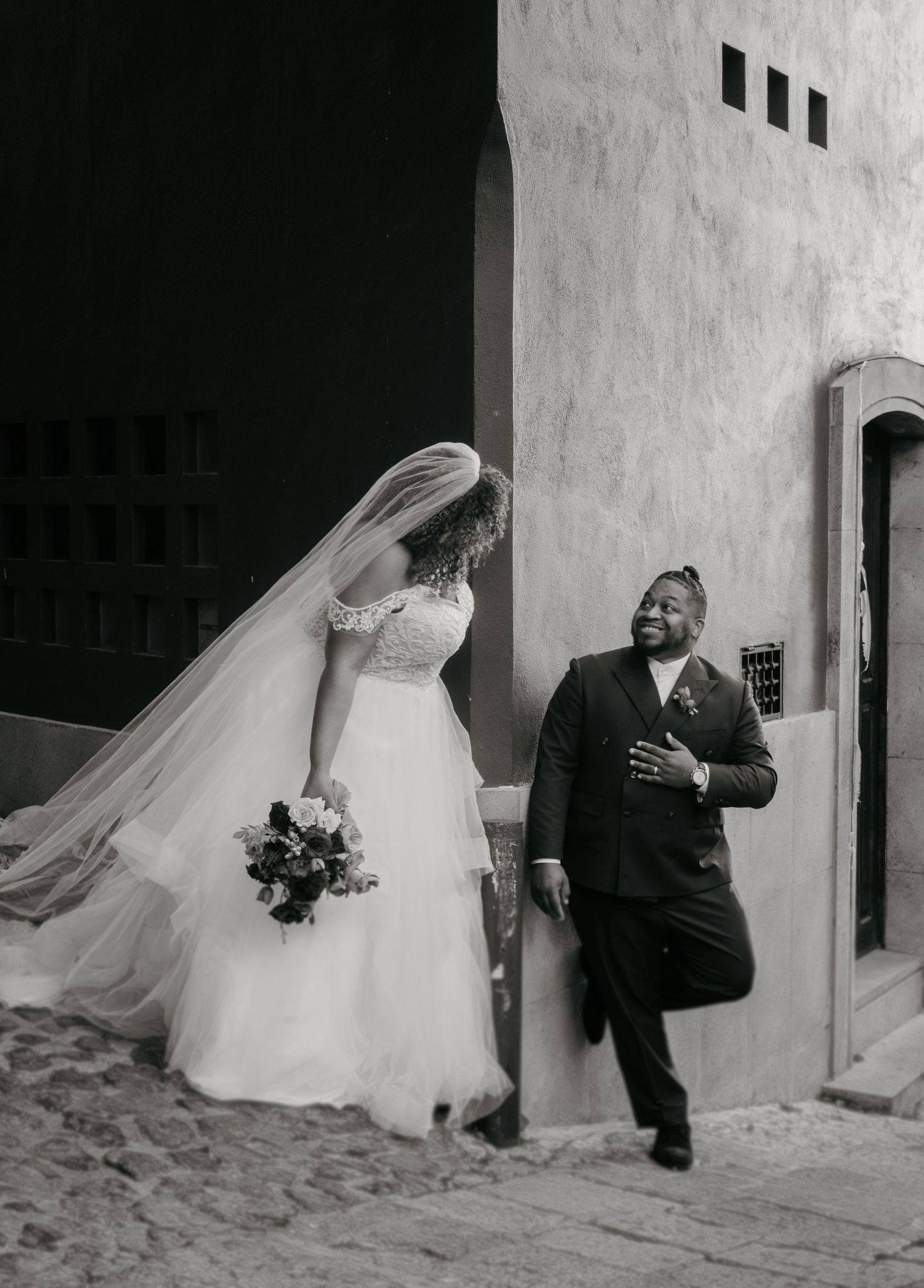 Black and white photo of a bride in a flowing gown and veil holding bouquet, smiling with groom in suit outdoors.