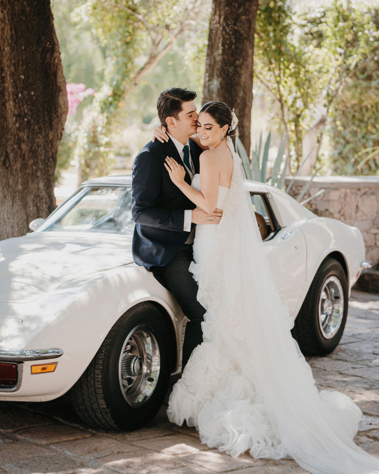 A bride and groom share a romantic kiss leaning against a classic white Corvette surrounded by lush trees.