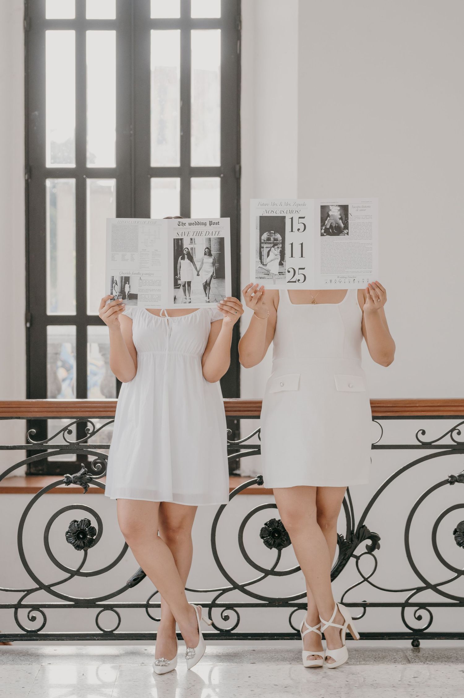 Two brides in white dresses hold wedding newspapers covering their faces on an elegant staircase.