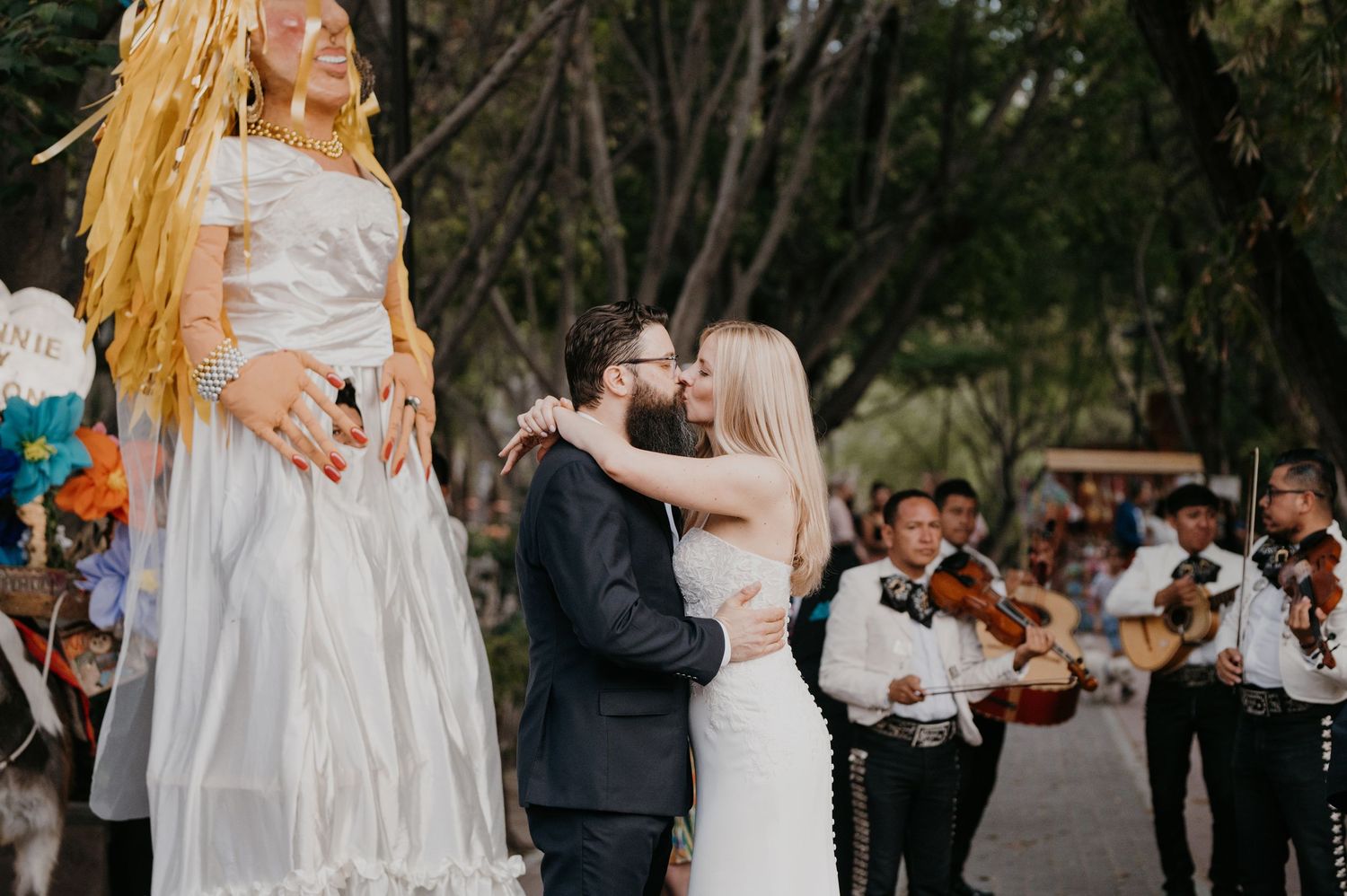 Bride and groom kiss surrounded by mariachi musicians and a colorful piñata at outdoor wedding.