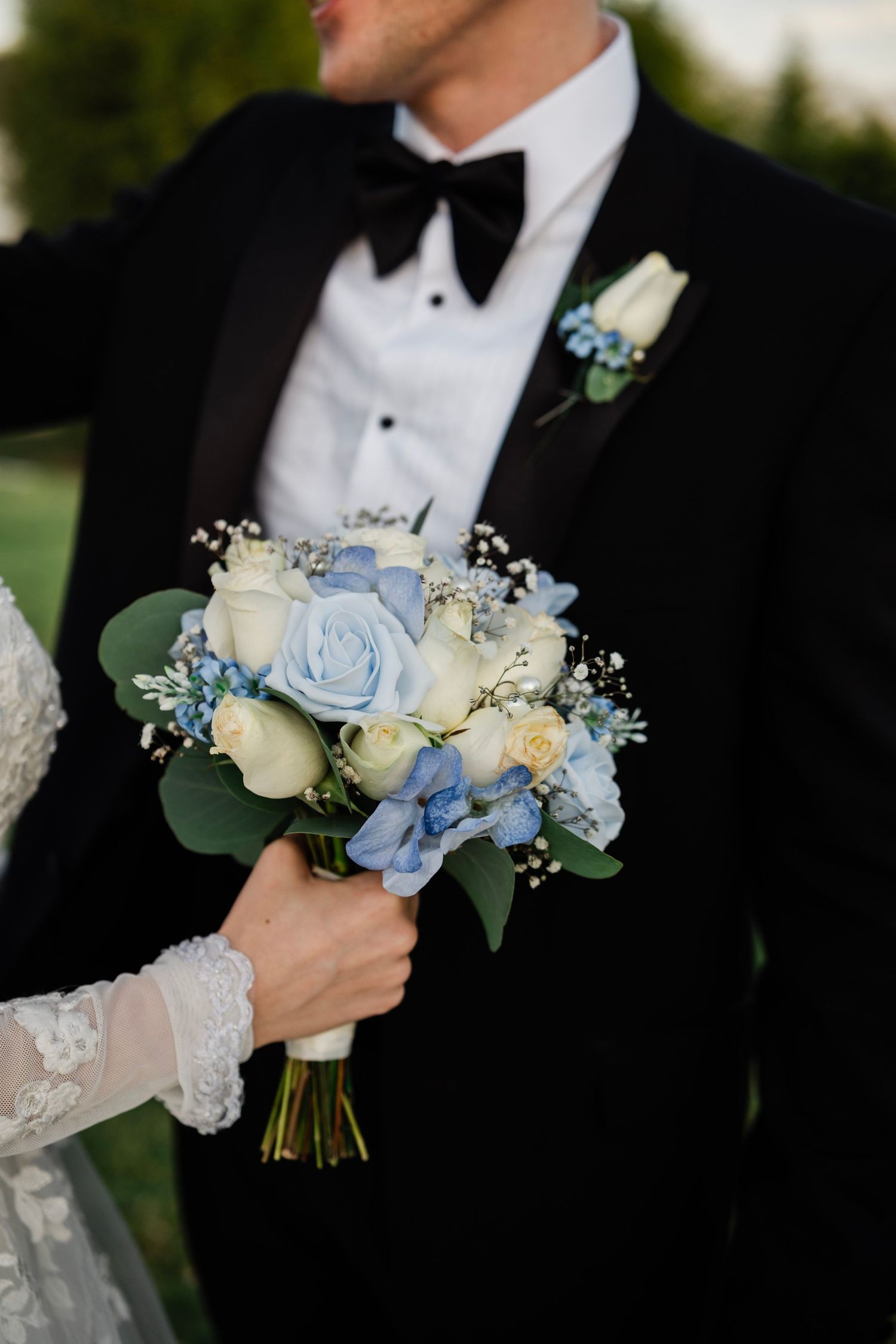Bride holding a blue and white wedding bouquet with roses and hydrangeas beside a groom in a black tuxedo.