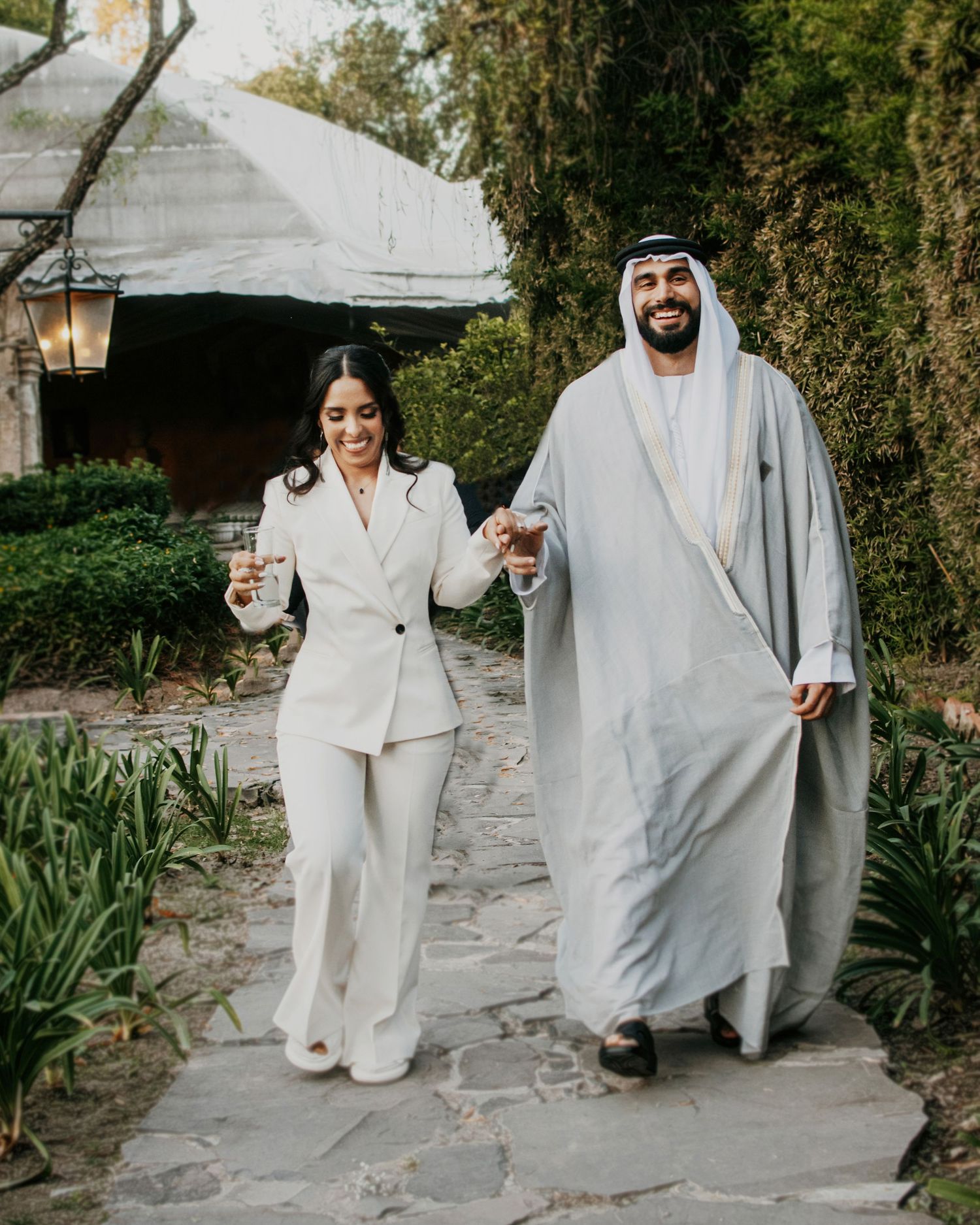 Couple in white suit and traditional UAE thobe walking hand in hand on stone path surrounded by lush greenery.