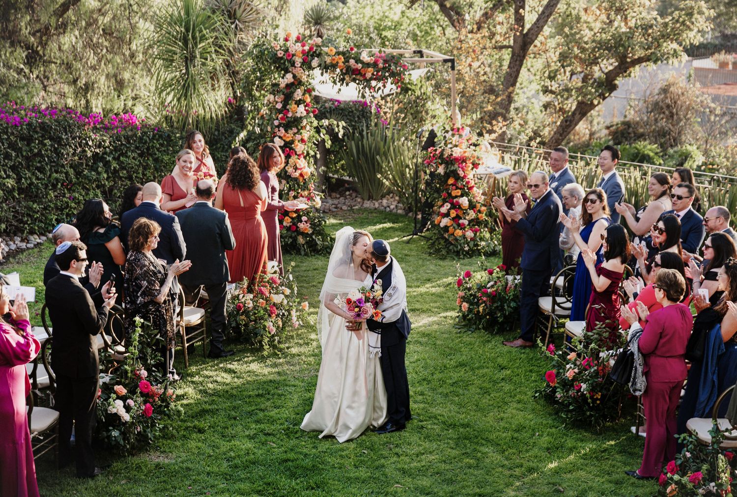 Bride and groom share first kiss at outdoor garden wedding ceremony with floral arch and cheering guests.