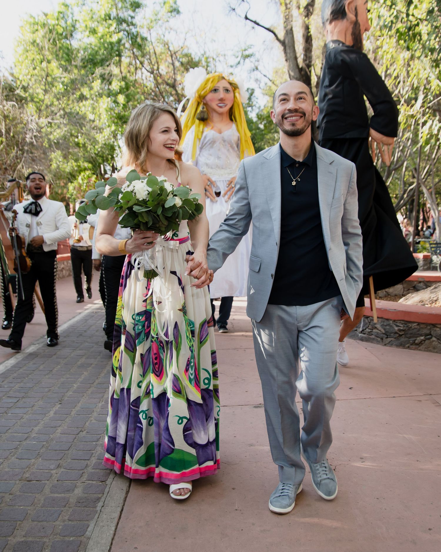 Happy couple walks hand-in-hand at outdoor wedding, bride in floral dress holding bouquet, groom in gray suit, guests behind.