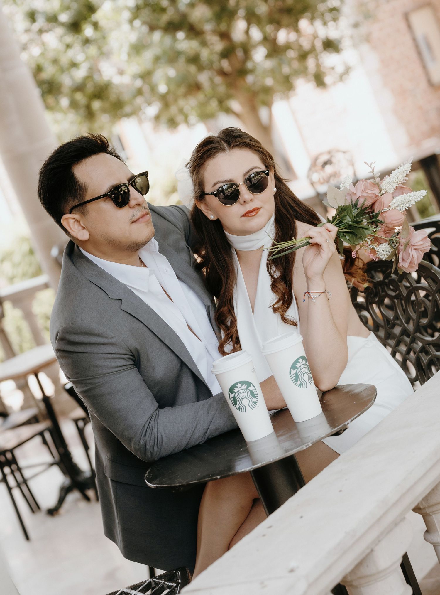 Stylish couple in sunglasses at Starbucks outdoor table, man in gray suit, woman in white dress holding pink bouquet.