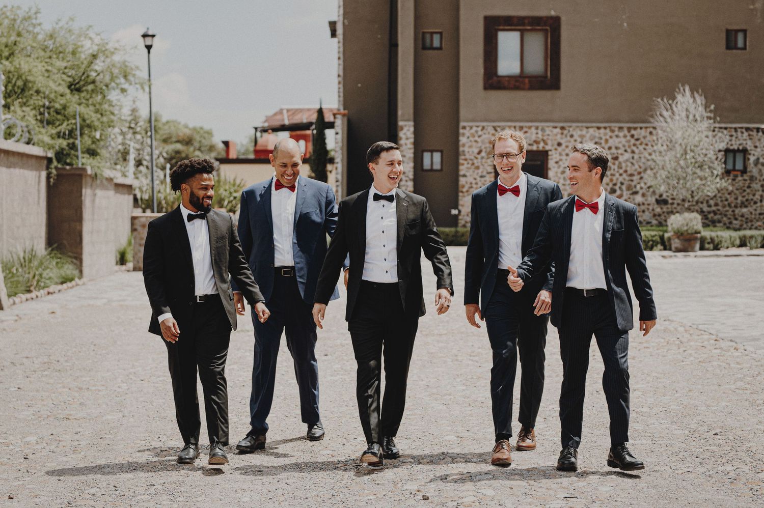 Five groomsmen in black tuxedos with bow ties walking outdoors on wedding day.