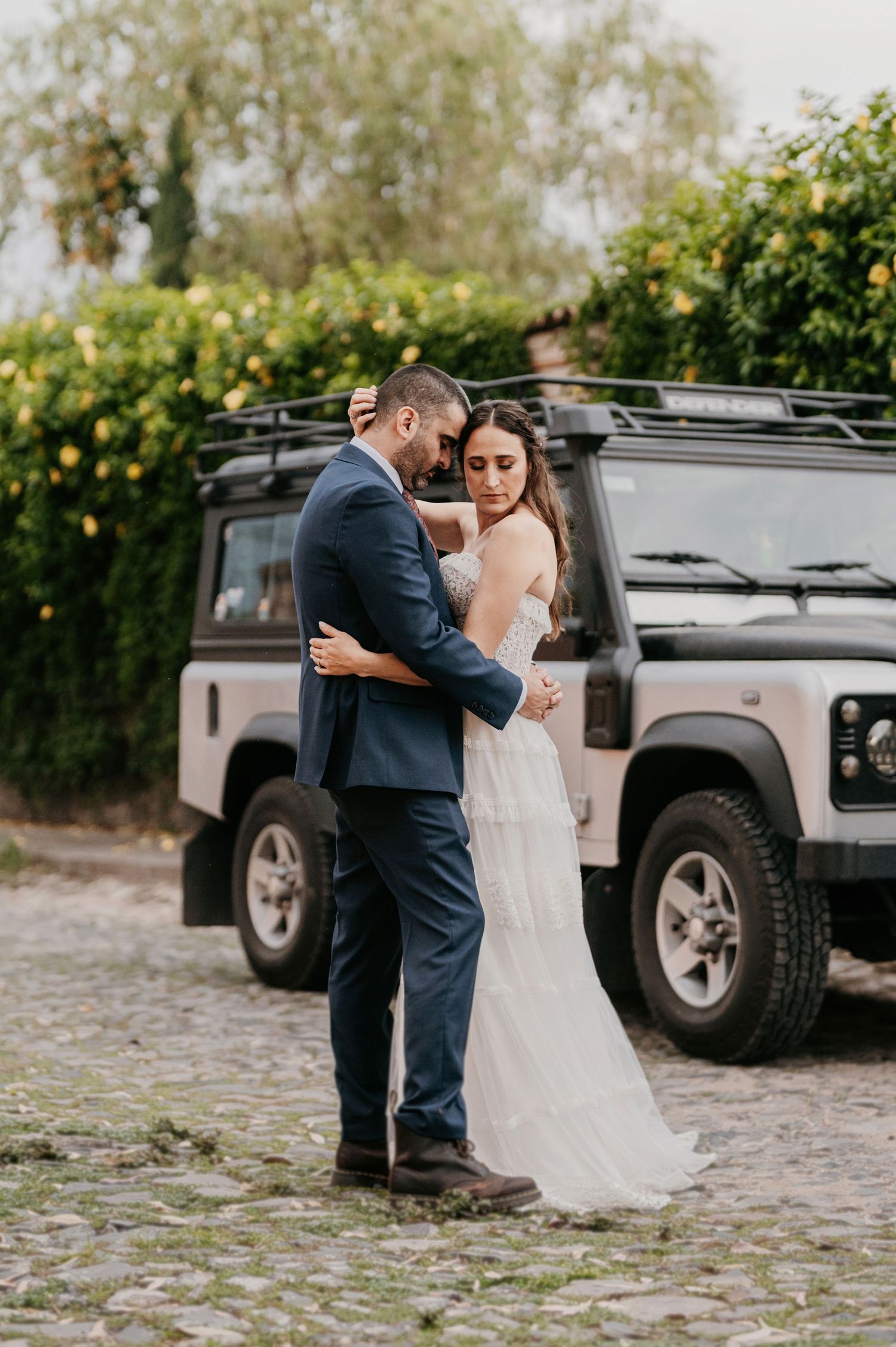 Bride and groom embracing in front of a black Land Rover Defender on a cobblestone path.