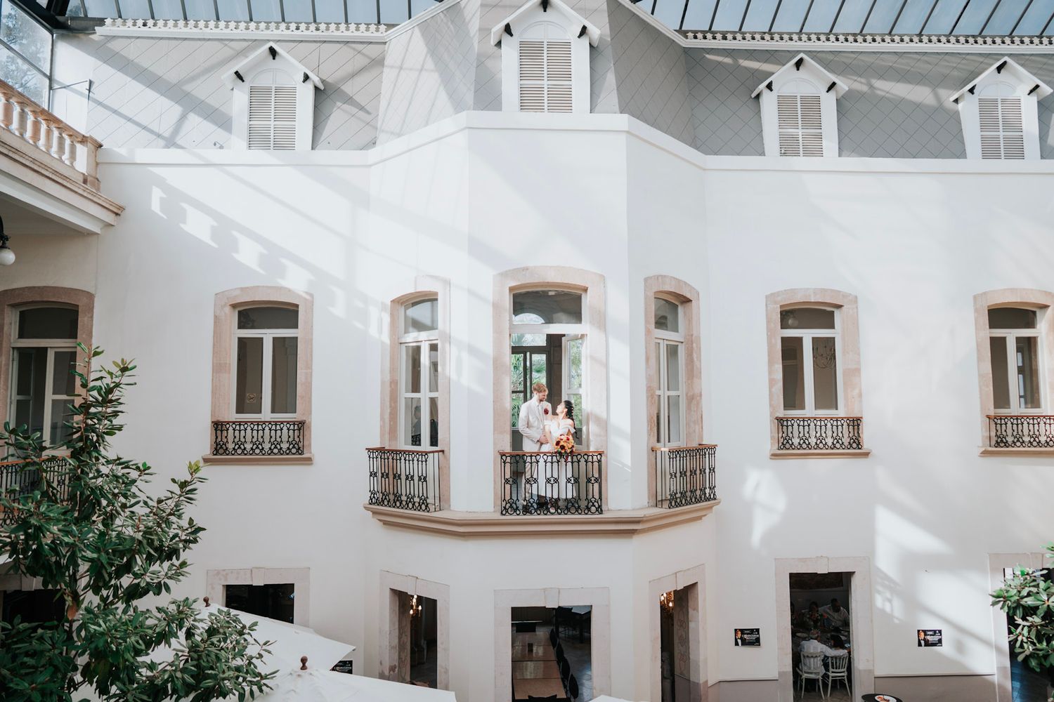 Wedding couple stands on elegant white building balcony with ornate ironwork in a sunlit courtyard.