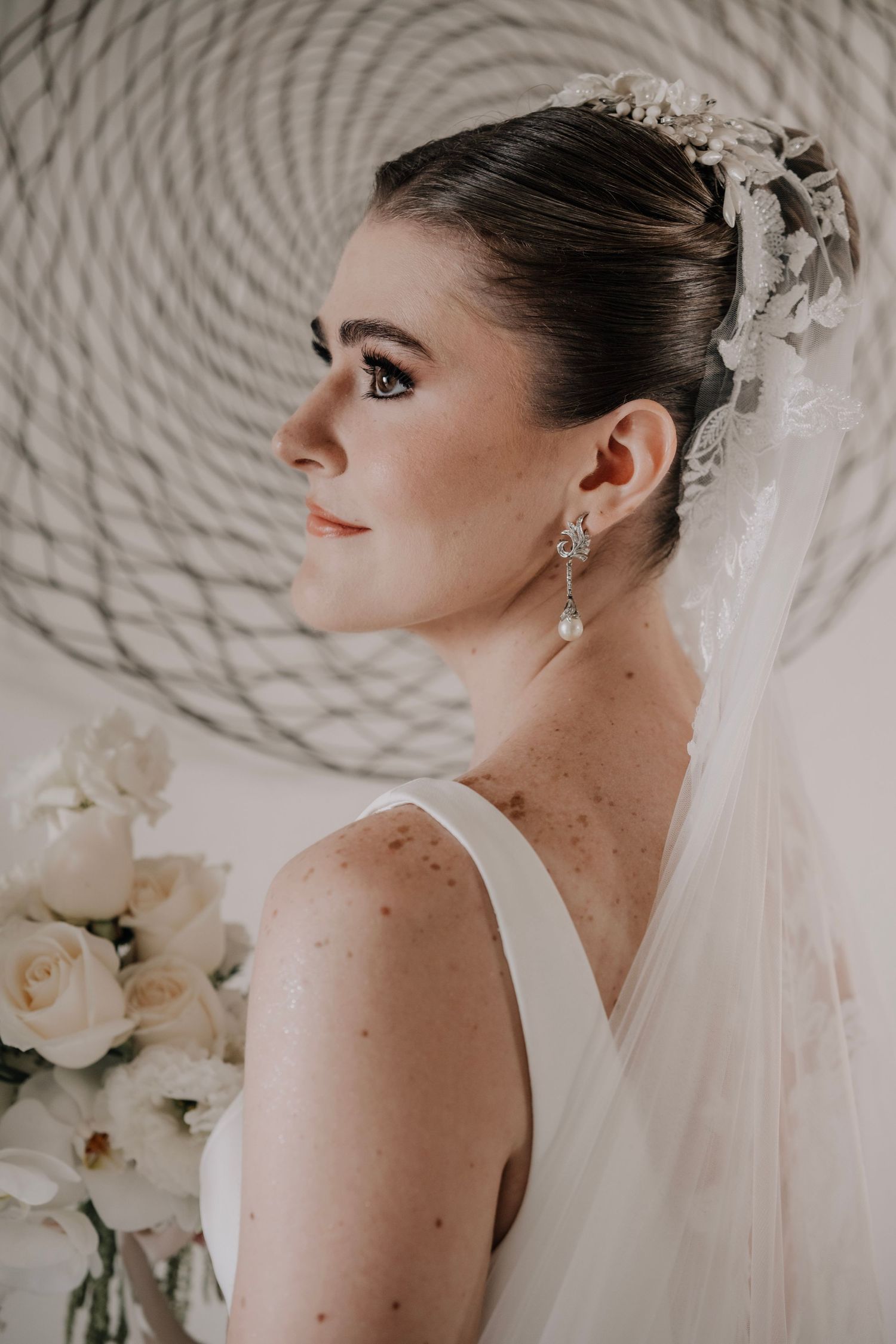 Elegant bride in profile wearing crystal drop earrings, floral headpiece, and sheer veil with white roses bouquet.