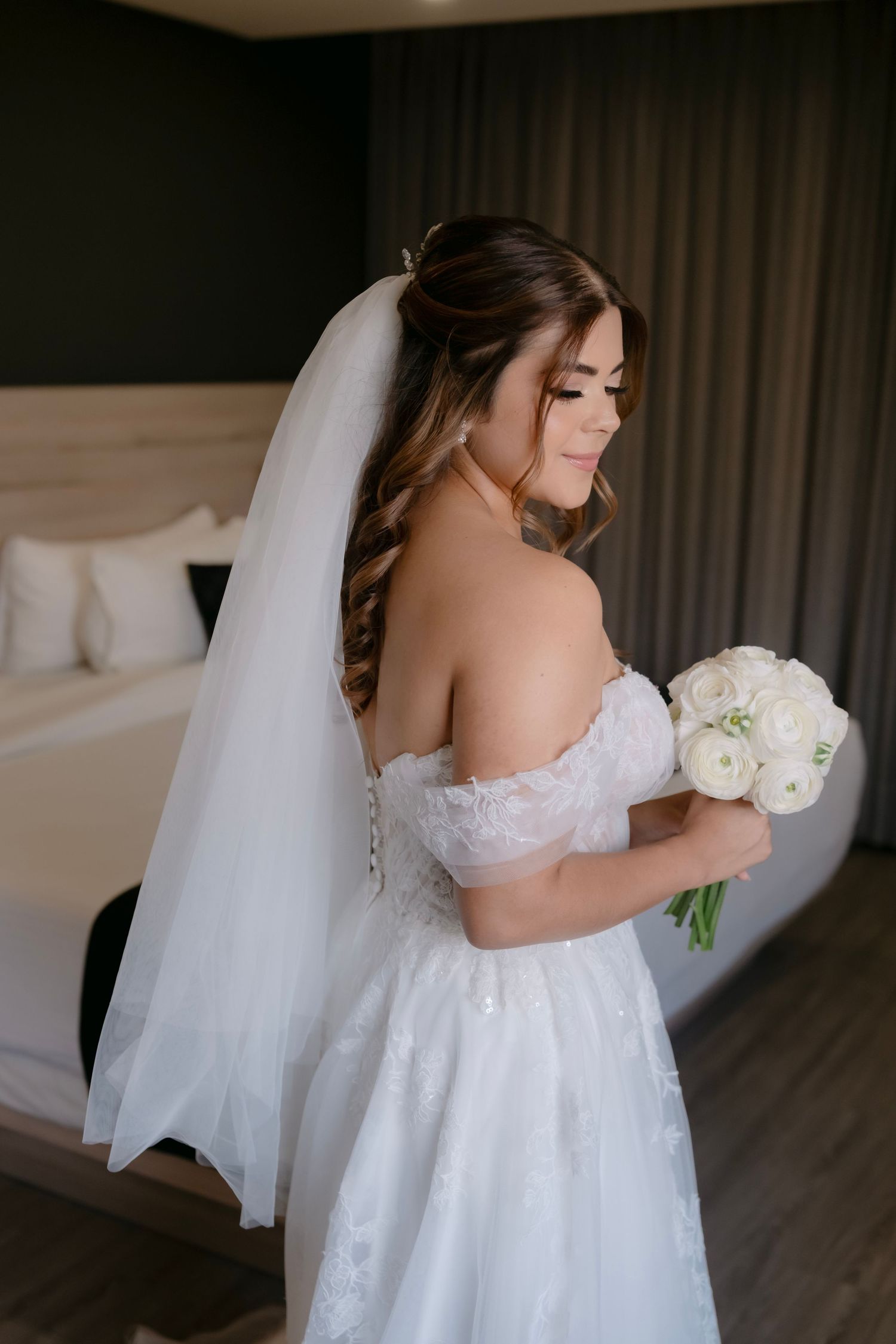 Bride in off-shoulder white lace gown holds bouquet of white roses, wearing veil in elegant hotel room.