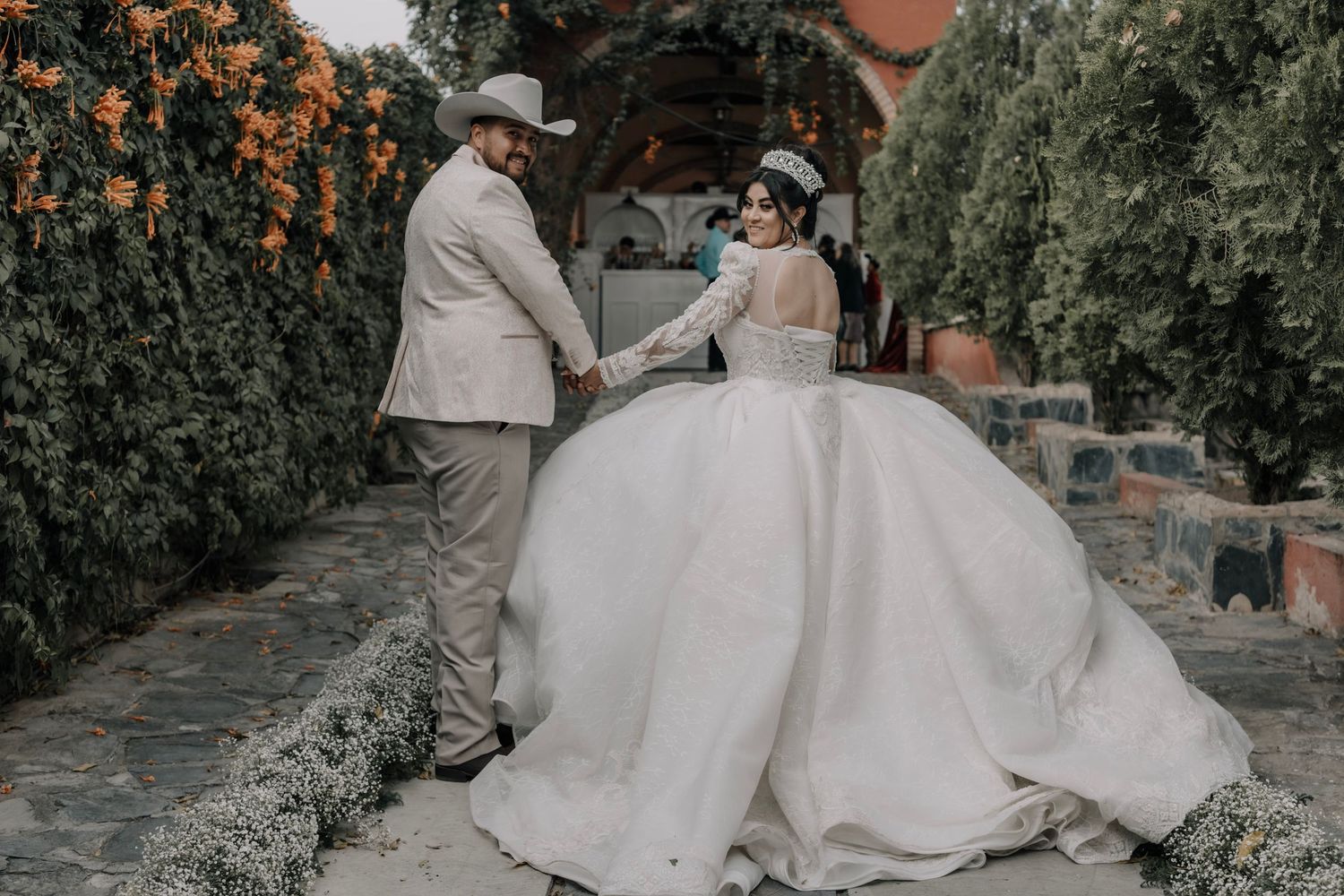 Bride in a white ball gown and groom in a tan suit hold hands in a lush garden wedding venue.