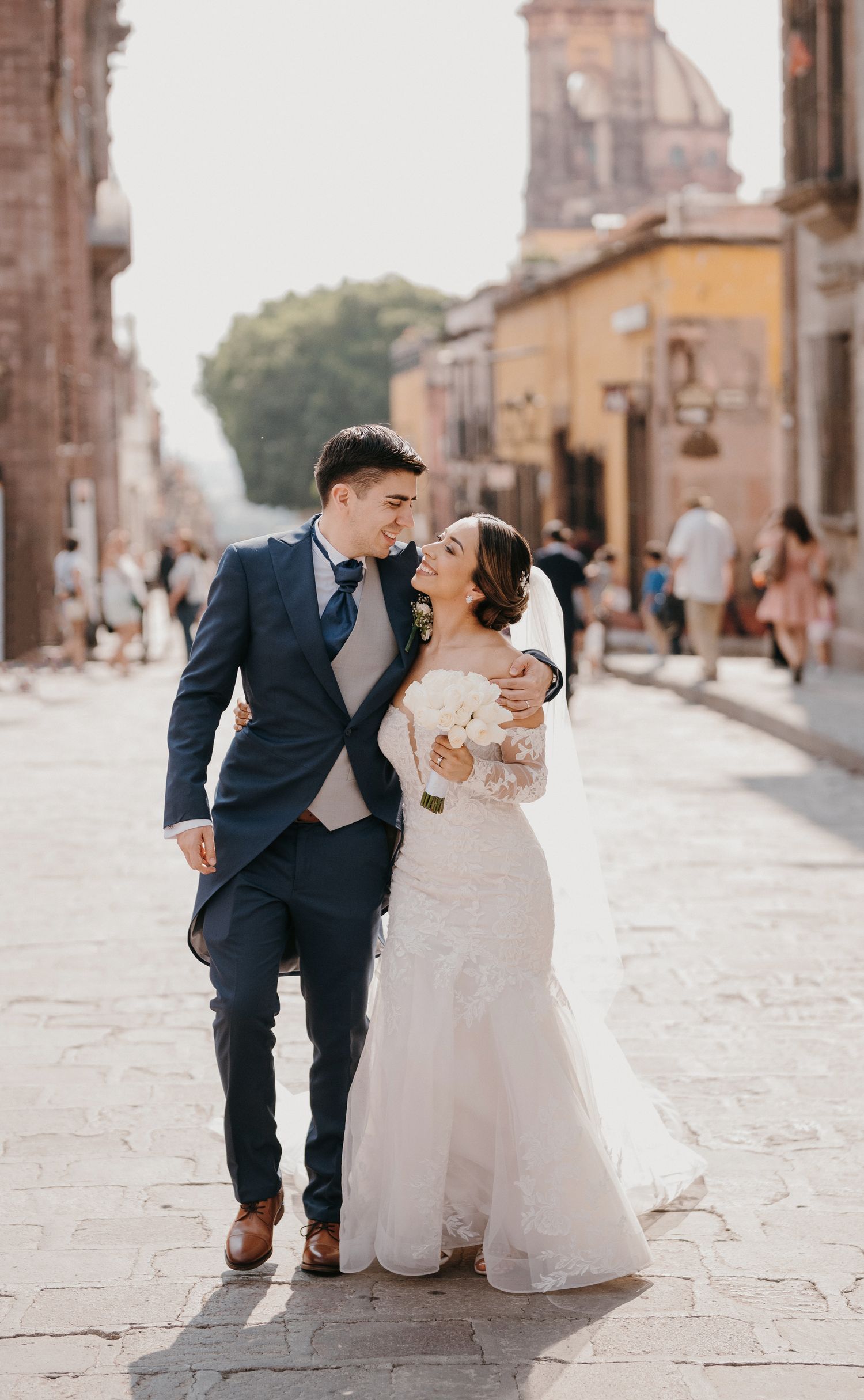 Newlyweds share a joyful moment walking cobblestone street, groom in navy suit, bride in lace gown.