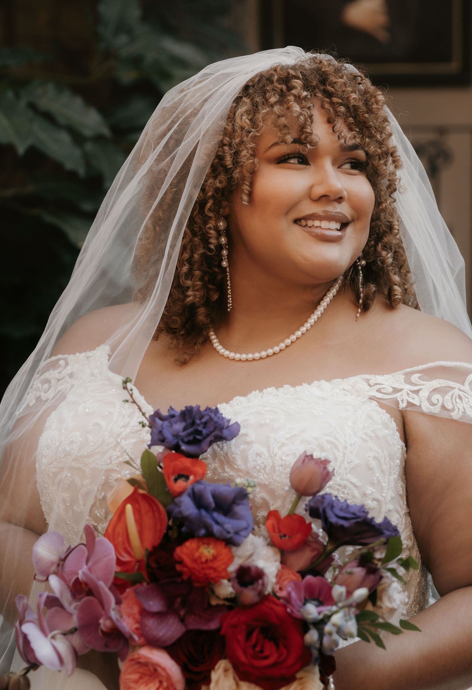 Beautiful bride with curly hair wearing a white lace gown, veil, and pearl necklace holding a colorful bouquet.