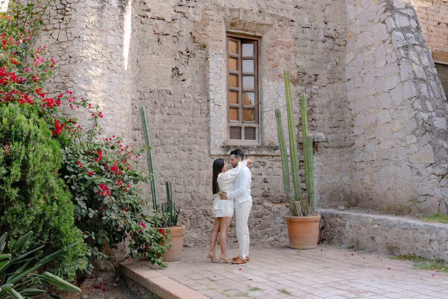 Couple embracing romantically in a rustic stone courtyard with cacti and red flowers.