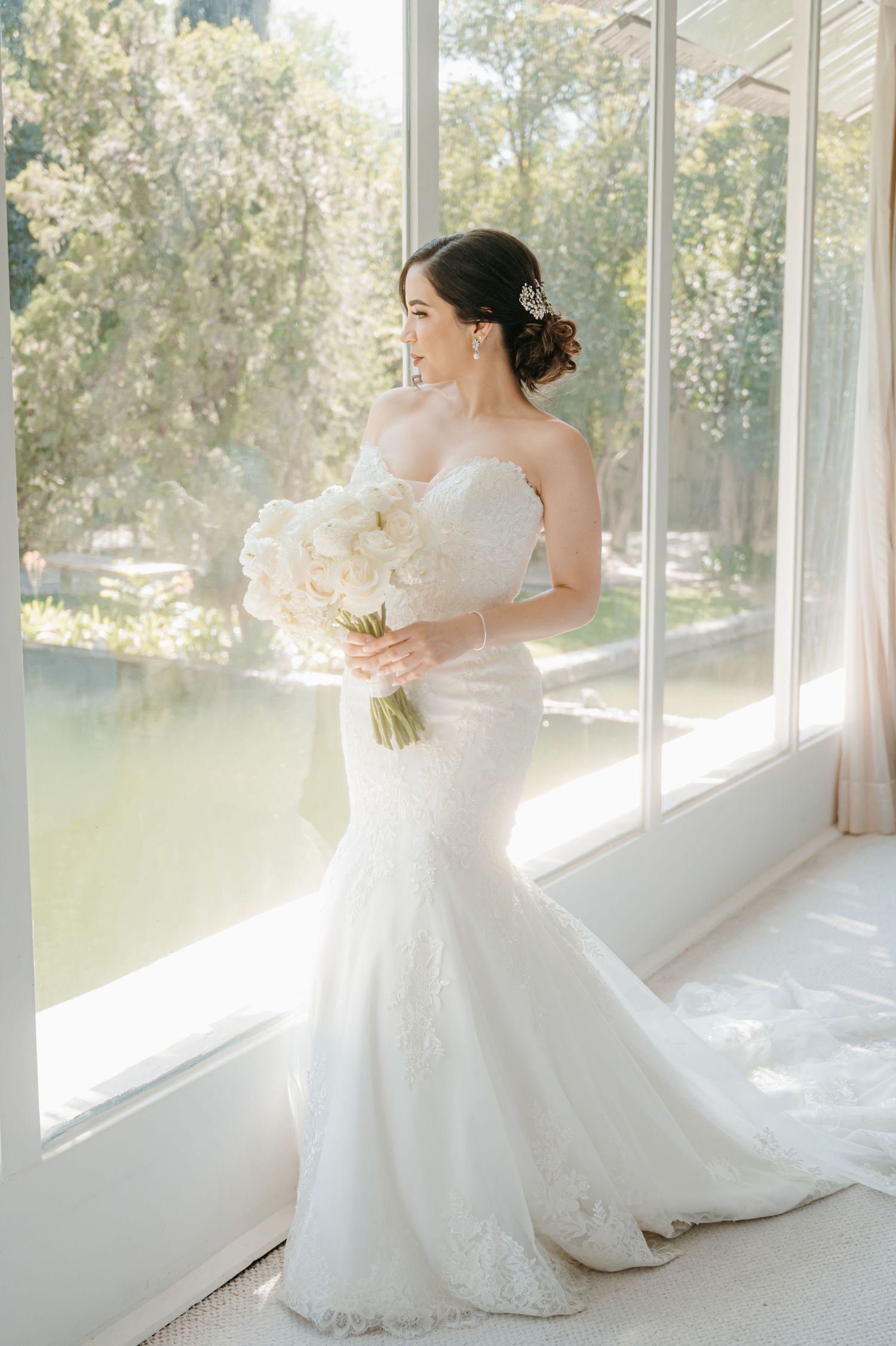 Bride in strapless lace mermaid gown holding white bouquet by sunlit garden window.