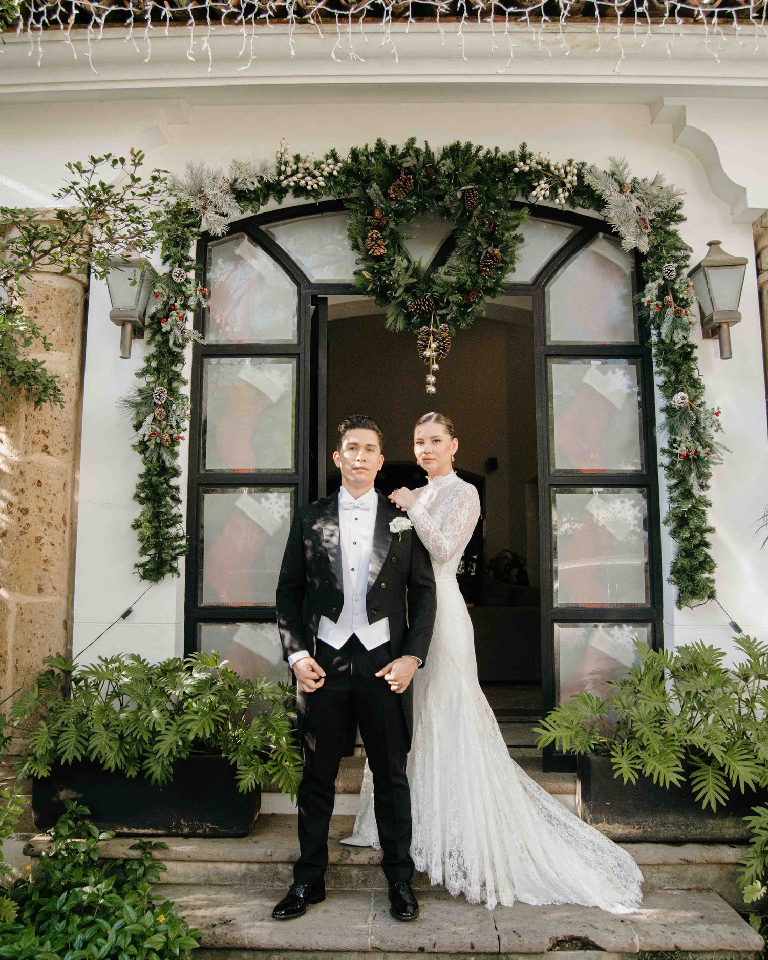 Bride and groom pose before a festive garland-adorned entrance with wreaths and greenery at a winter wedding.