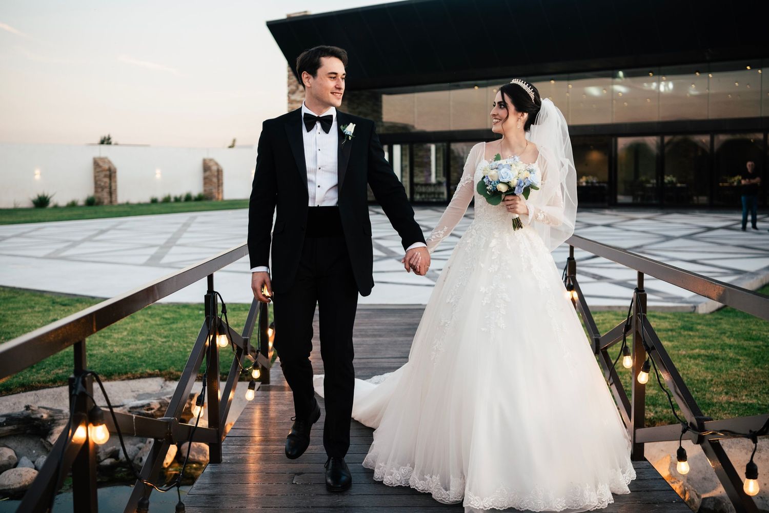 Bride and groom hold hands walking down outdoor steps at dusk, she in white ballgown, he in black tuxedo.