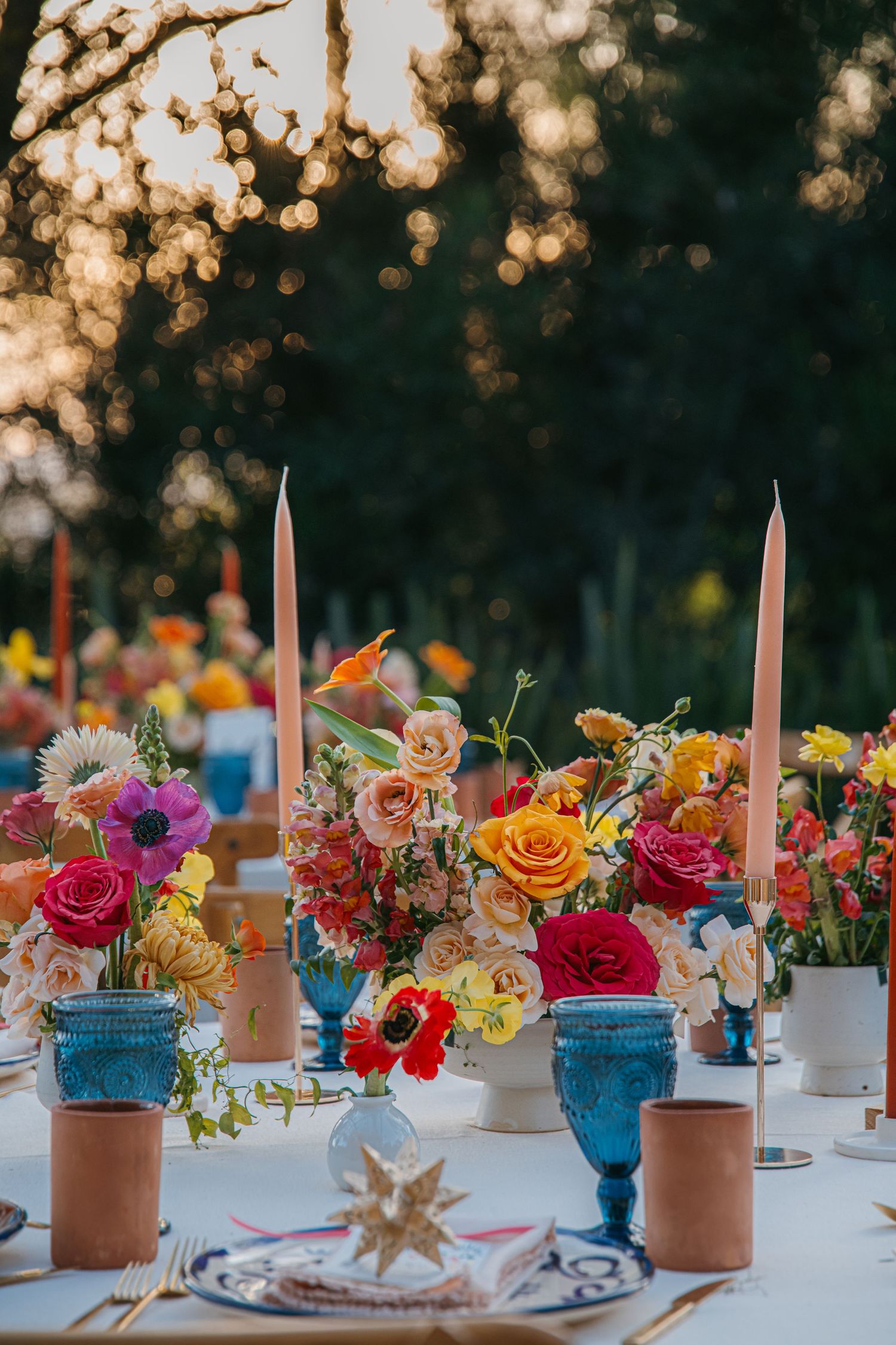Colorful outdoor wedding tablescape with vibrant floral centerpieces, pink taper candles, blue goblets, and terracotta vases at sunset.